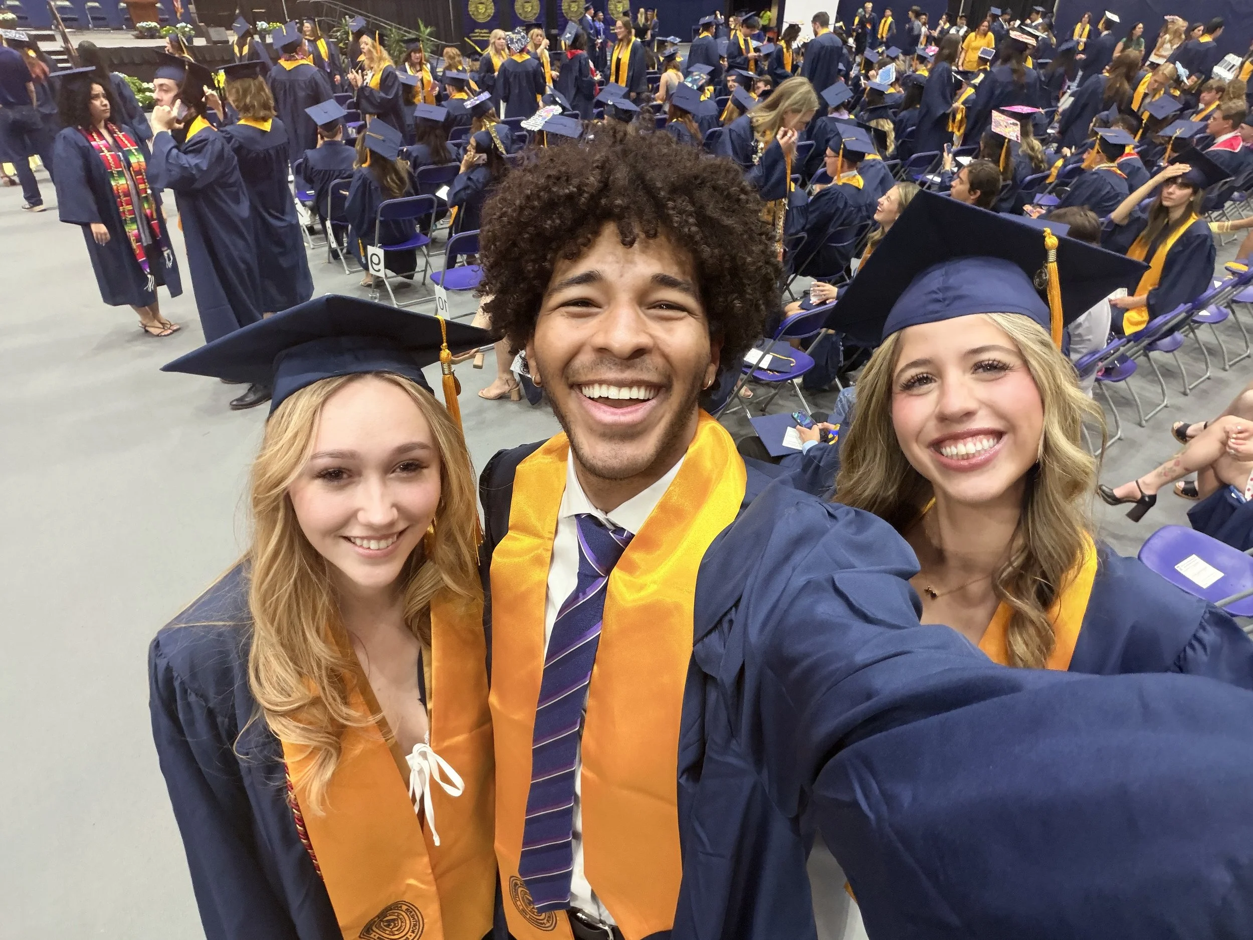 Group of graduates in caps and gowns taking a selfie at their graduation ceremony, with many graduates seated in the background.