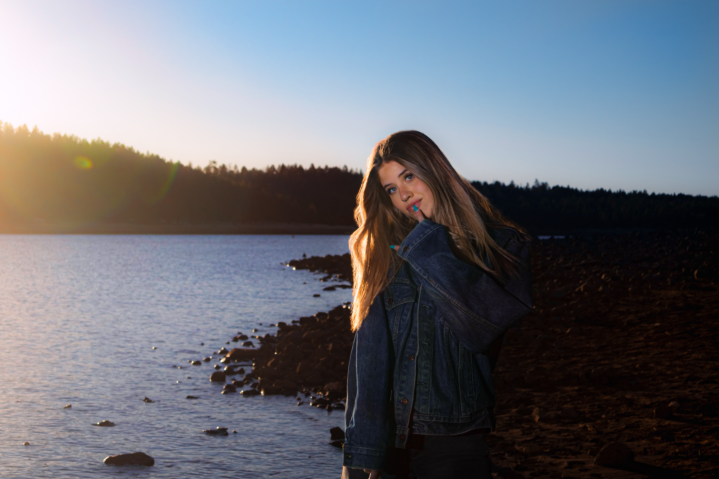 A young woman with long hair and painted nails posing by a lakeshore at sunset, wearing a denim jacket.