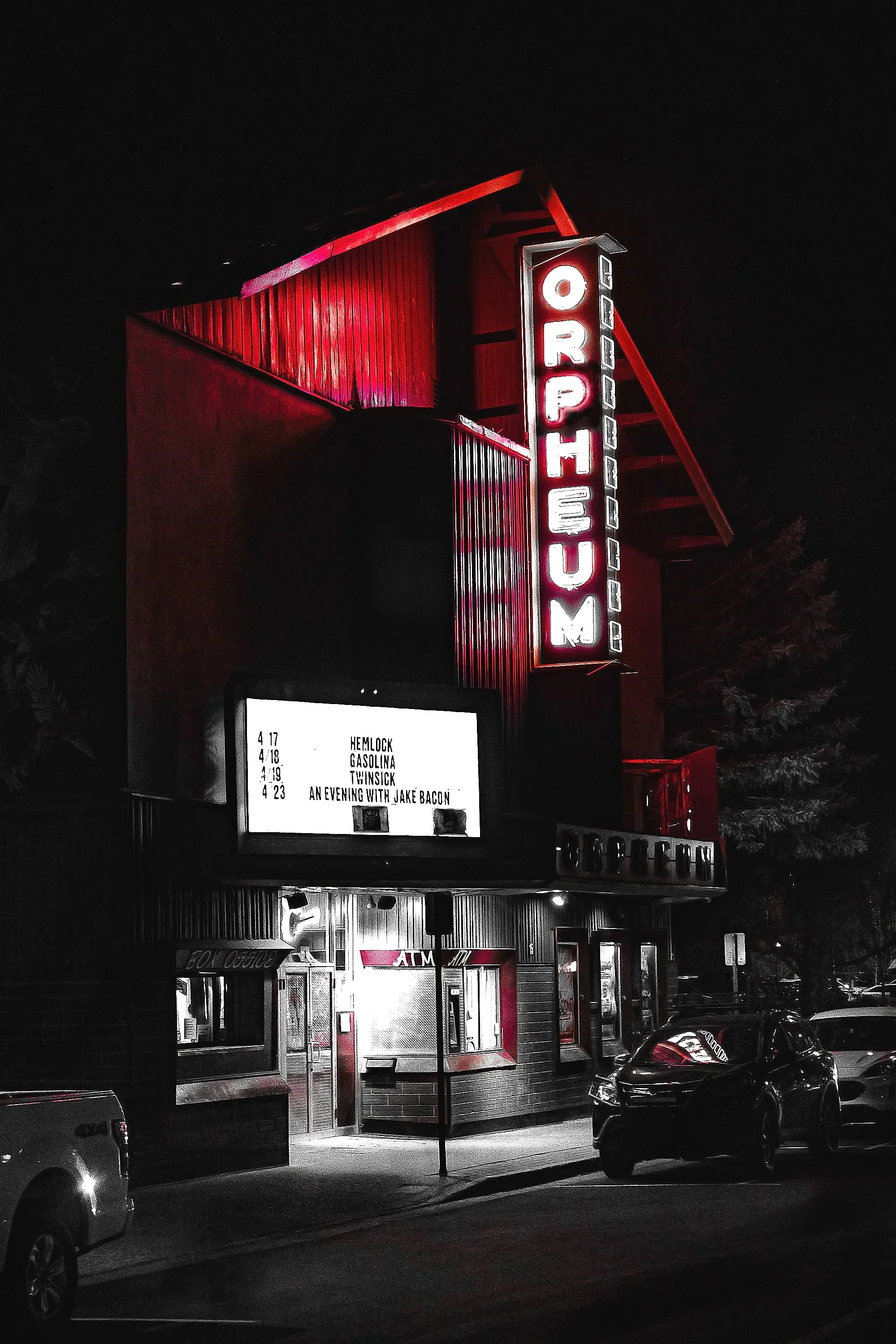 A photo of a red and black movie theater at night with illuminated vertical sign displaying the theater's name. The front of the theater has an entrance with a small awning, illuminated signs, and a parking lot with a few cars. There is also a digita