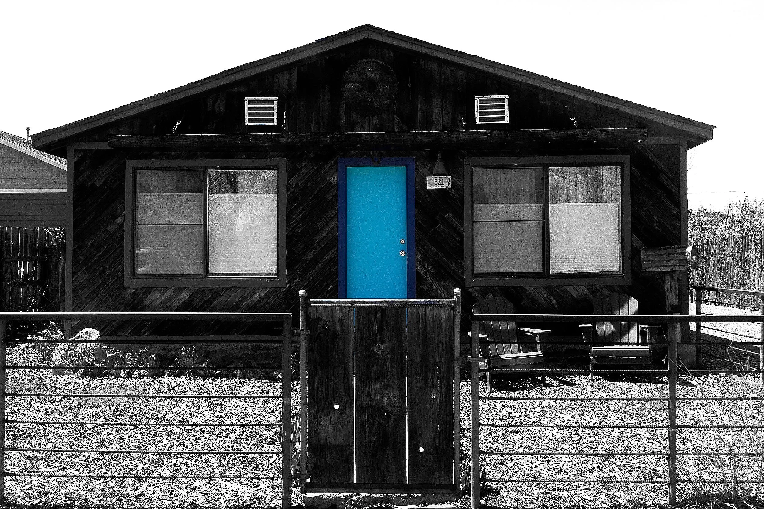 Front view of a small house with a bright blue door, two large windows, a fenced yard, and a black skull decoration near the roof, captured in black and white.
