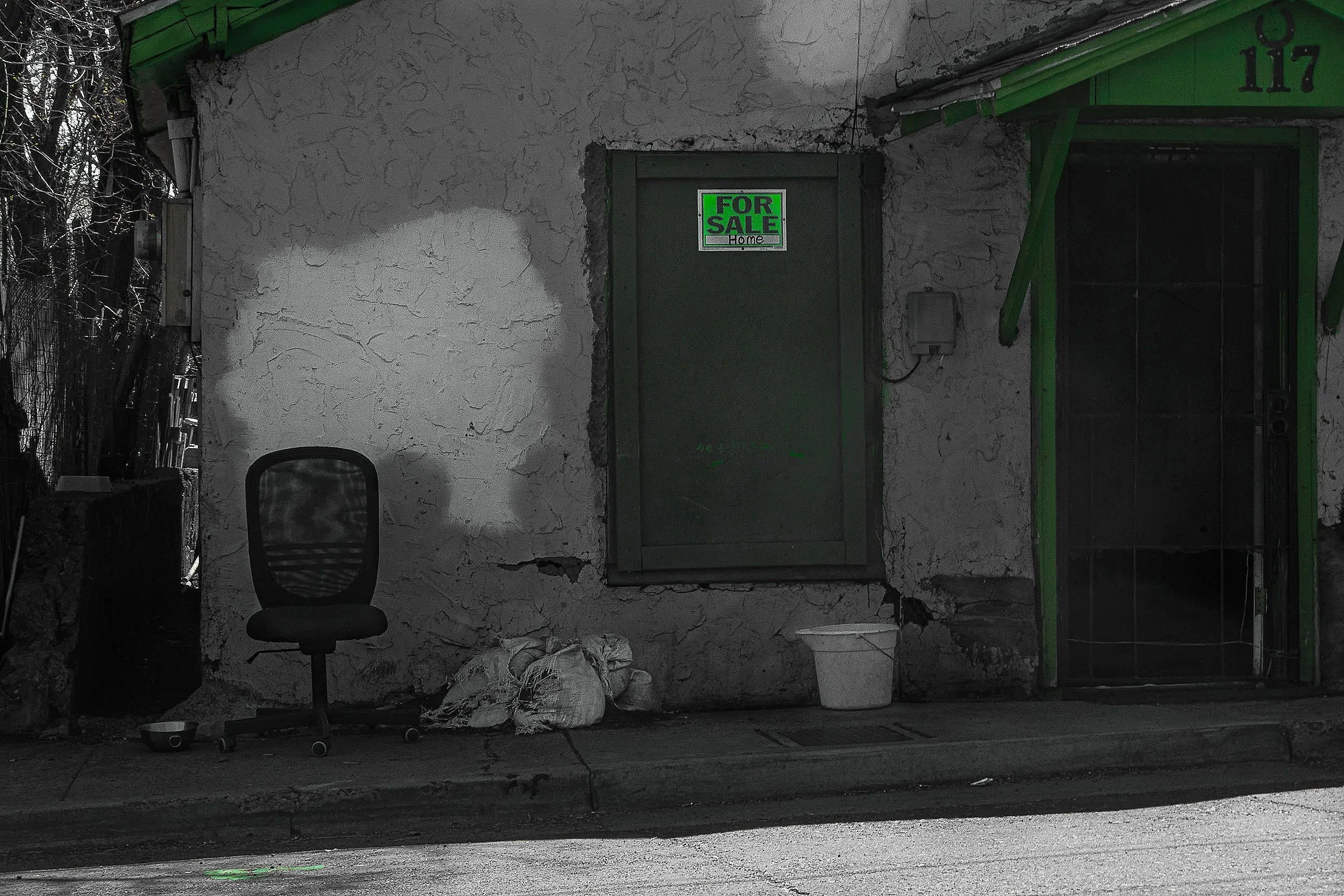 The image depicts an aged, textured white wall with a small door in the center. There is a 'For Sale' sign on the door. To the right, part of a green awning is visible above a window. In front of the wall, there is a black office chair, and to the le