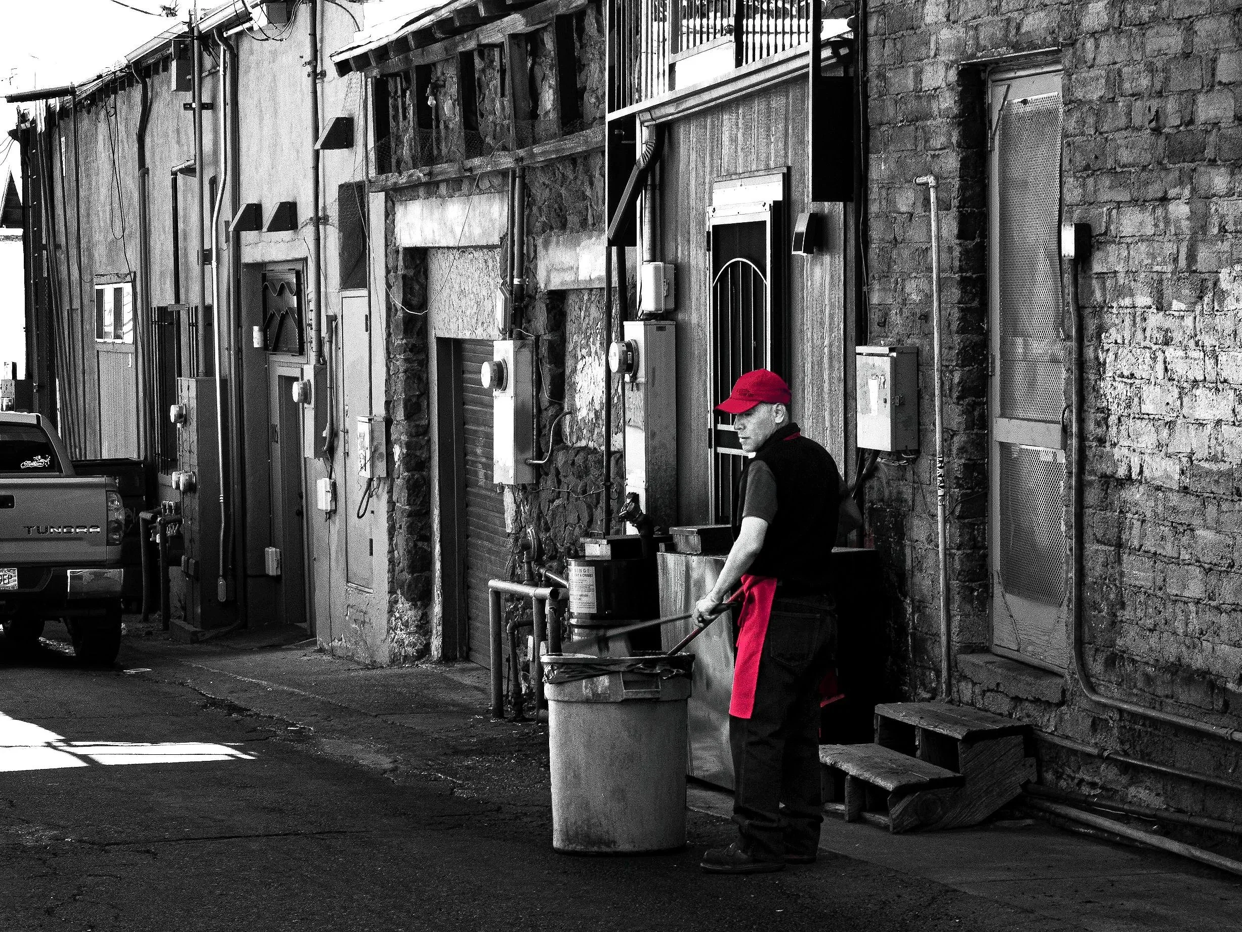 A man with a red cap and apron stands on a city sidewalk next to a large trash bin, with a stone wall and building wall behind him. The scene is black and white, but the man's cap and apron are in color.