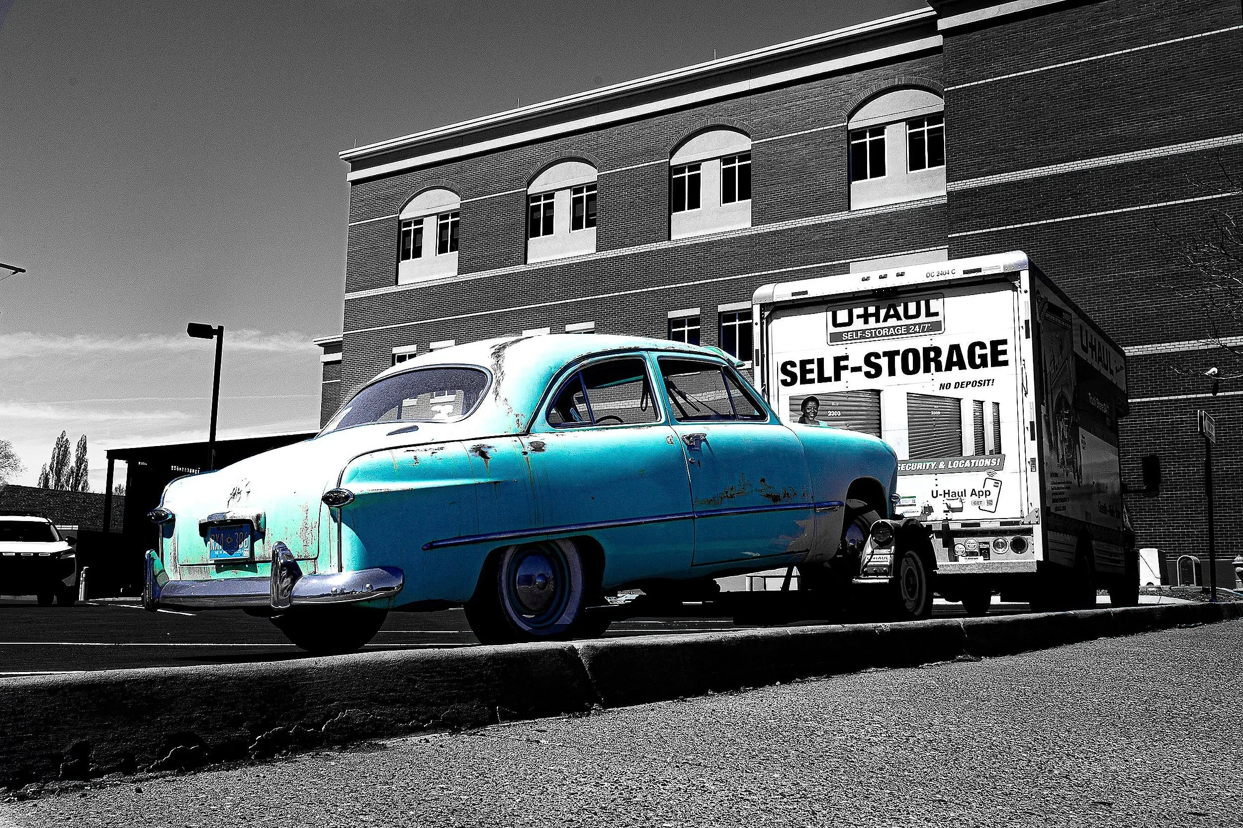 An old, rusted vintage car in blue and white, parked in a lot next to a U-Haul self-storage truck, with a brick building in the background.