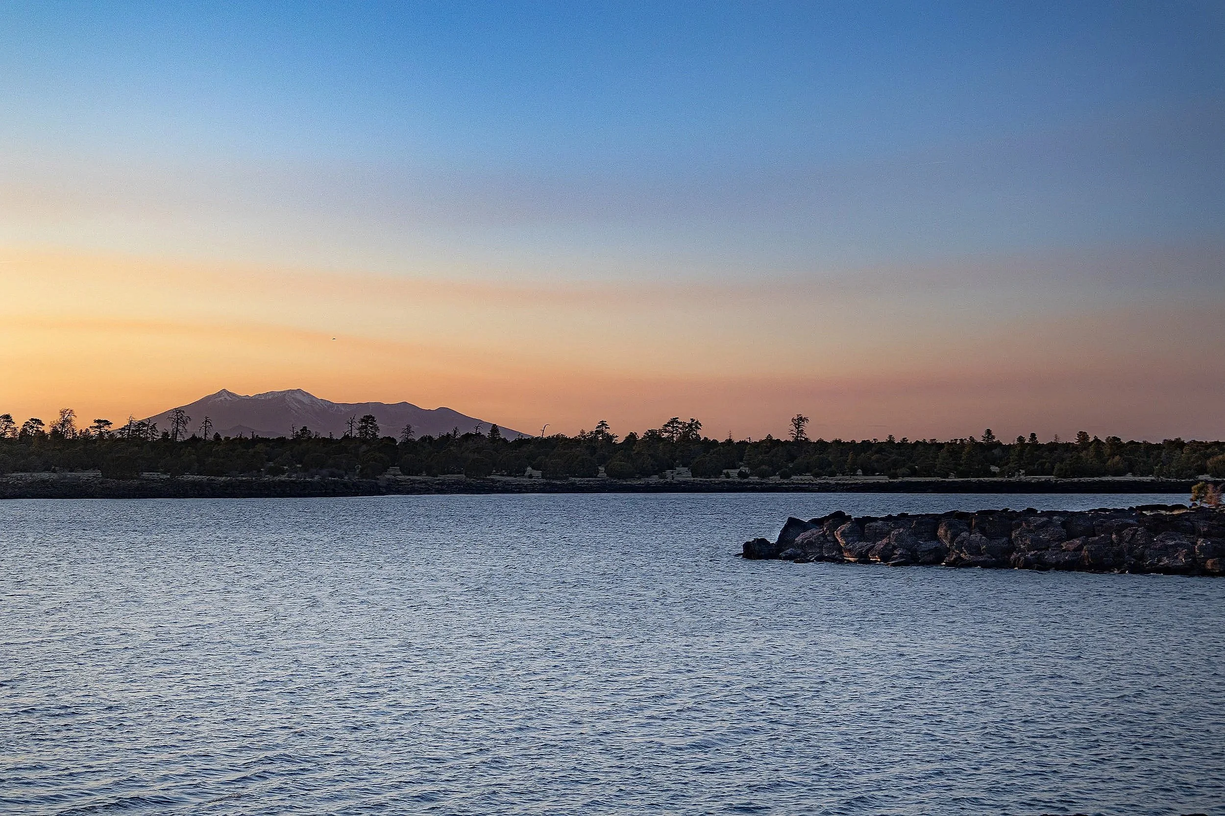 A scenic landscape with a calm body of water in the foreground, a forested shoreline in the middle, and mountains in the background under a clear sky with some pink and orange hues from the setting sun.