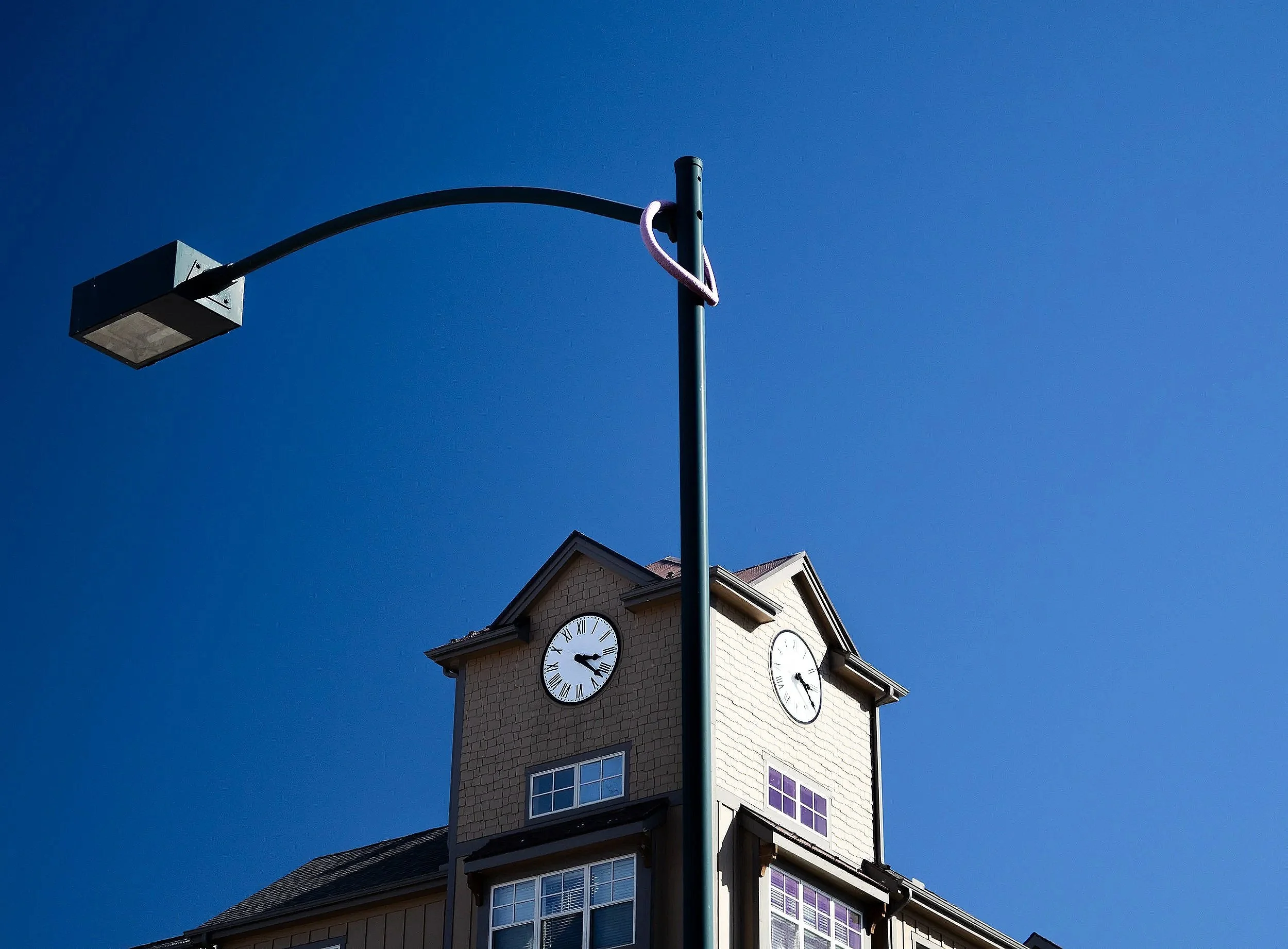 A clock tower with multiple clock faces showing different times, a street lamp with a pink cord wrapped around it, and a house with a tiled roof and windows, all set against a clear blue sky.