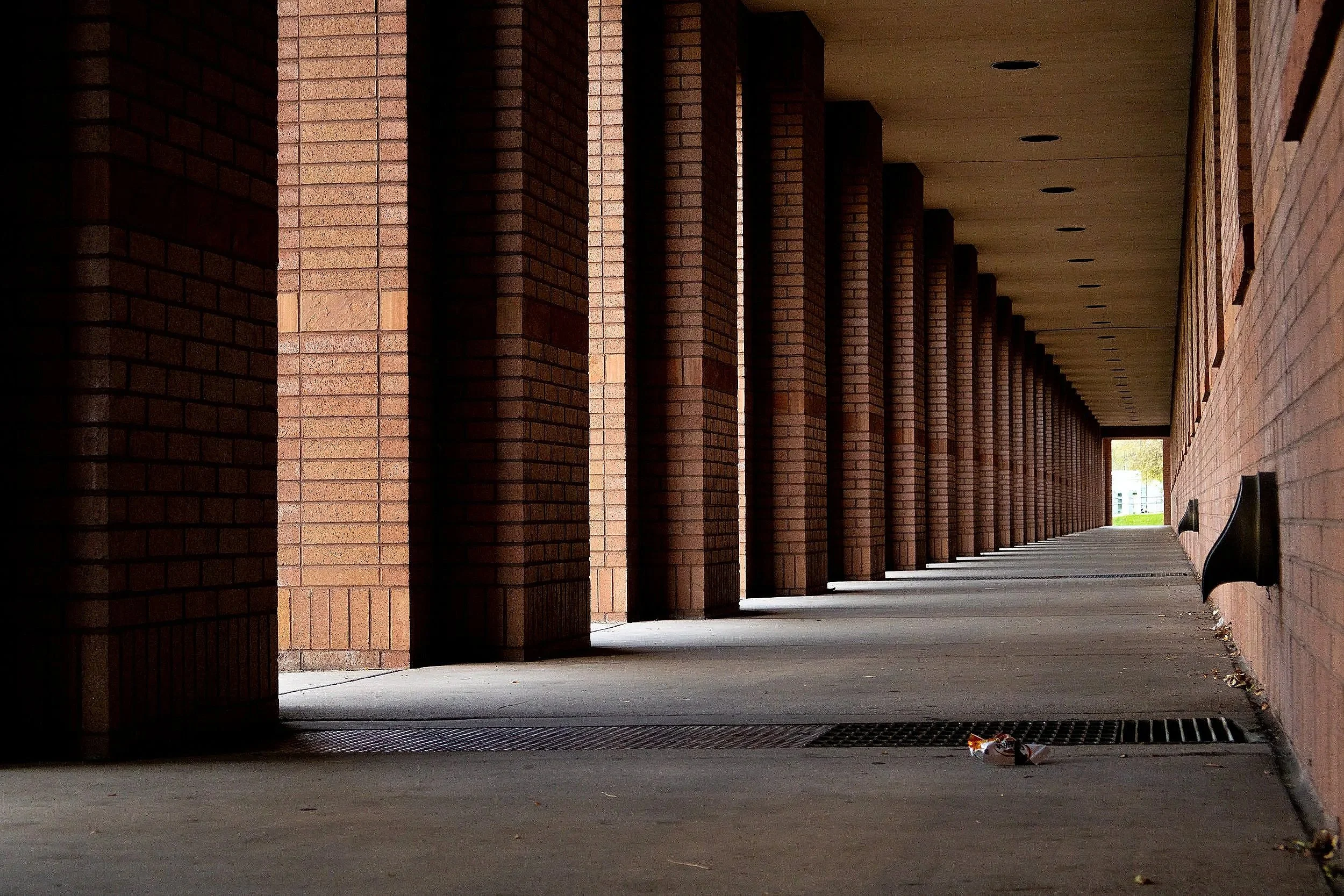 A long walkway with brick columns on the left side and a brick wall on the right side, leading to an opening with trees outside.