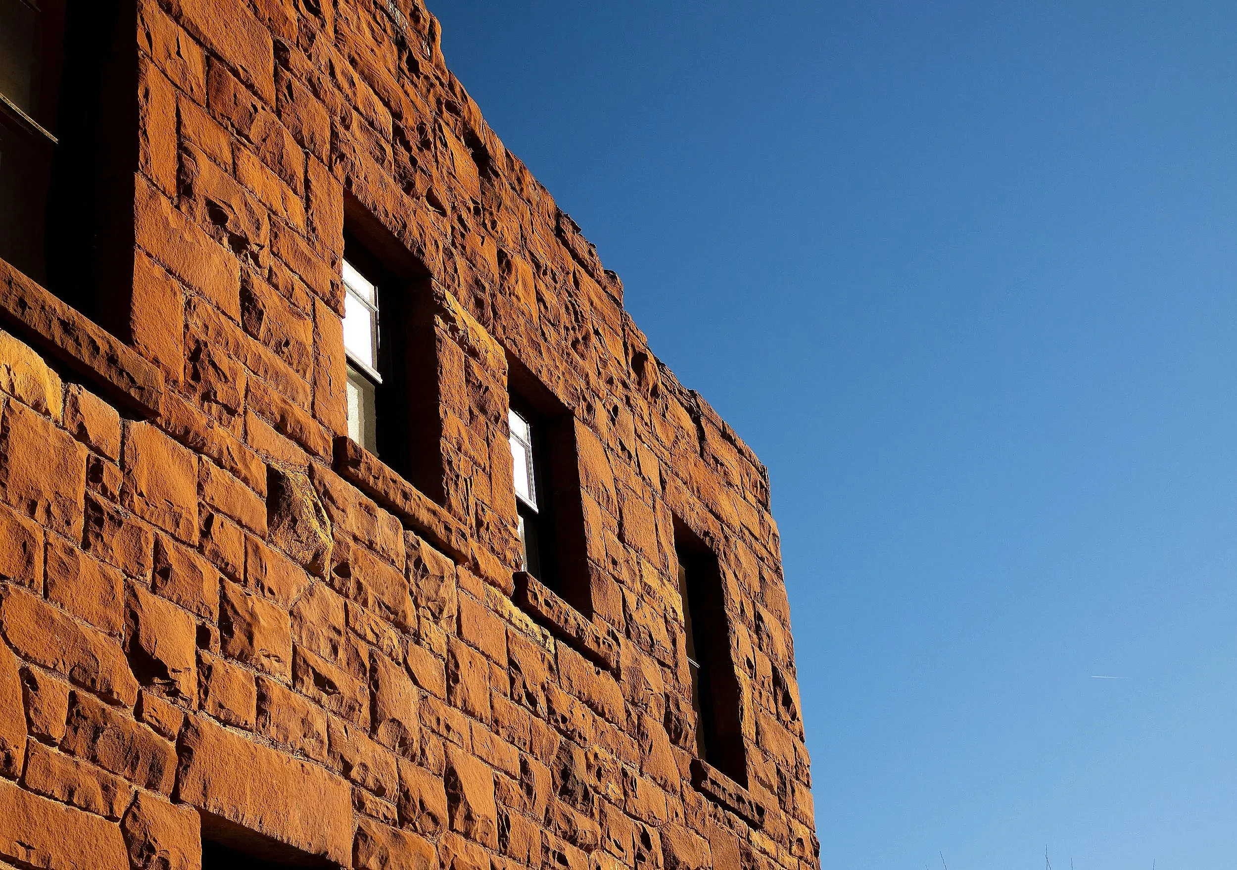 The photo shows a close-up view of a building made of reddish-brown bricks, with a clear blue sky in the background. The building has several small rectangular windows.