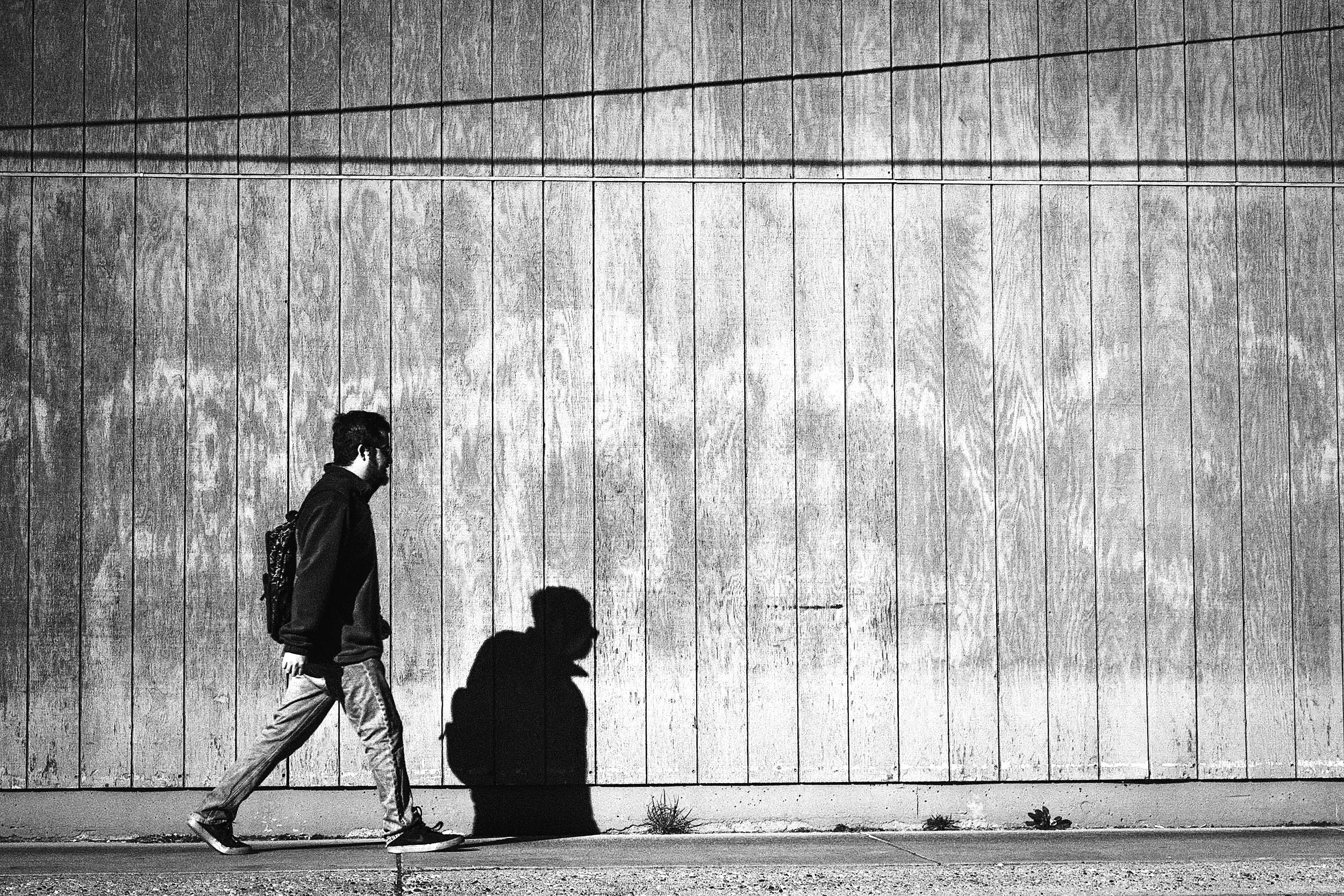 A person walking along a sidewalk casts a shadow on a large, textured concrete wall.