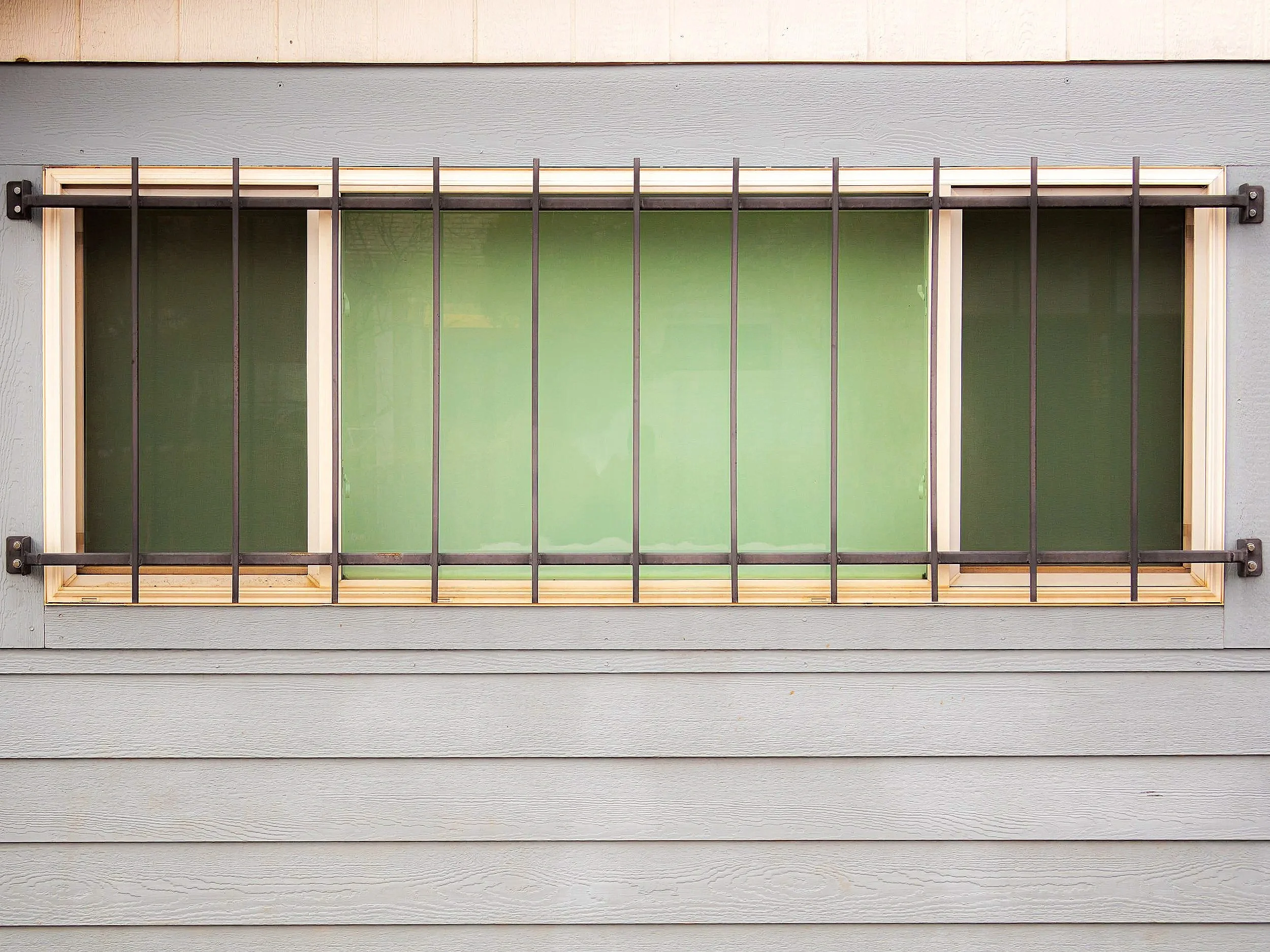 A window with green-tinted glass panes protected by black metal bars, set into a gray wooden wall with horizontal siding.
