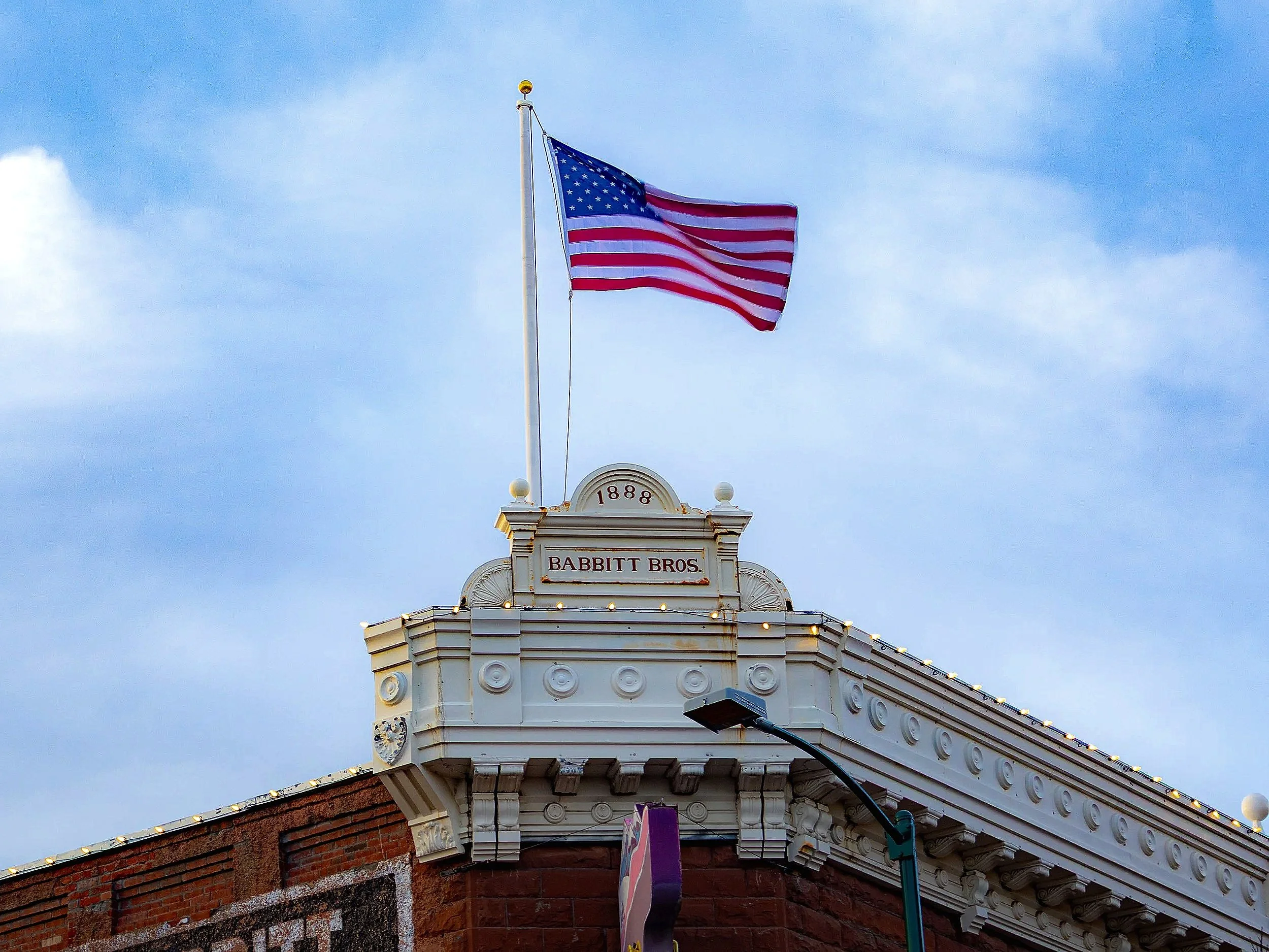 An American flag flying on a flagpole atop a historical building with the year 1888 and the words Babbitt Bros. on a sign, against a partly cloudy sky.