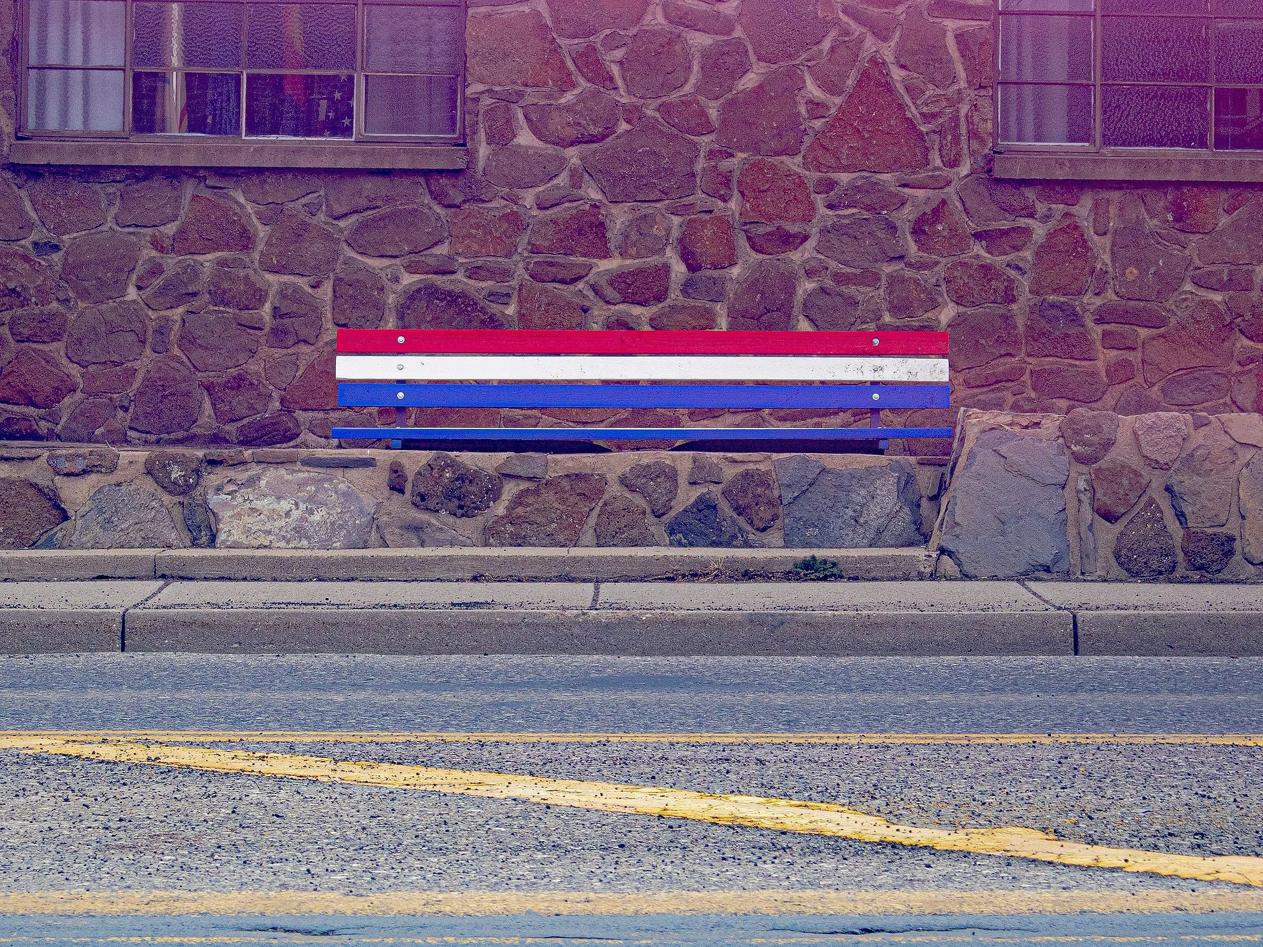 A stone building with two windows and a bench painted in red, white, and blue composite, placed in front of the building. The ground has a yellow parking lot line.