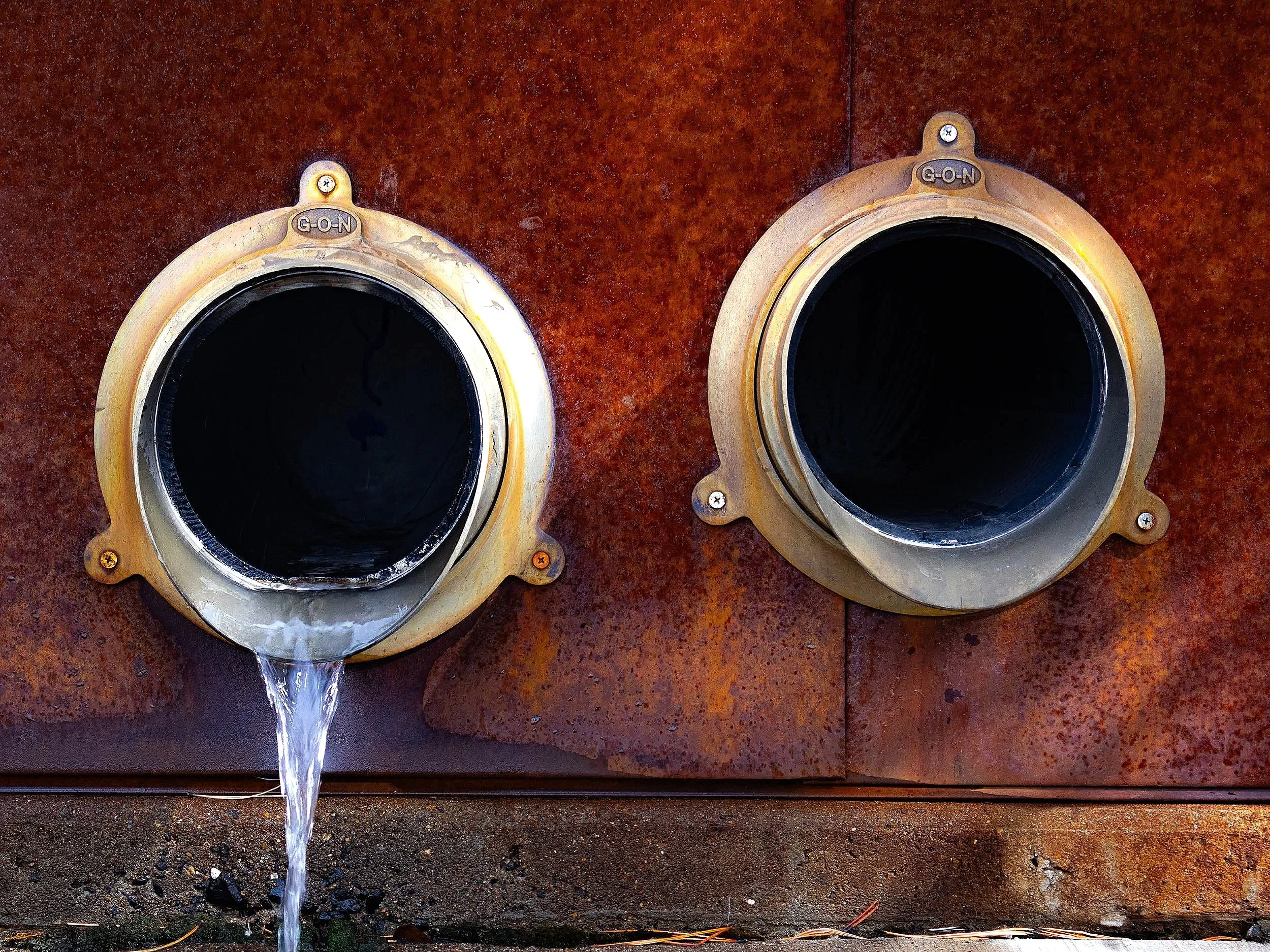 Two large circular metal fixtures attached to a rusty surface, with water flowing from the bottom of each fixture onto the surface below.