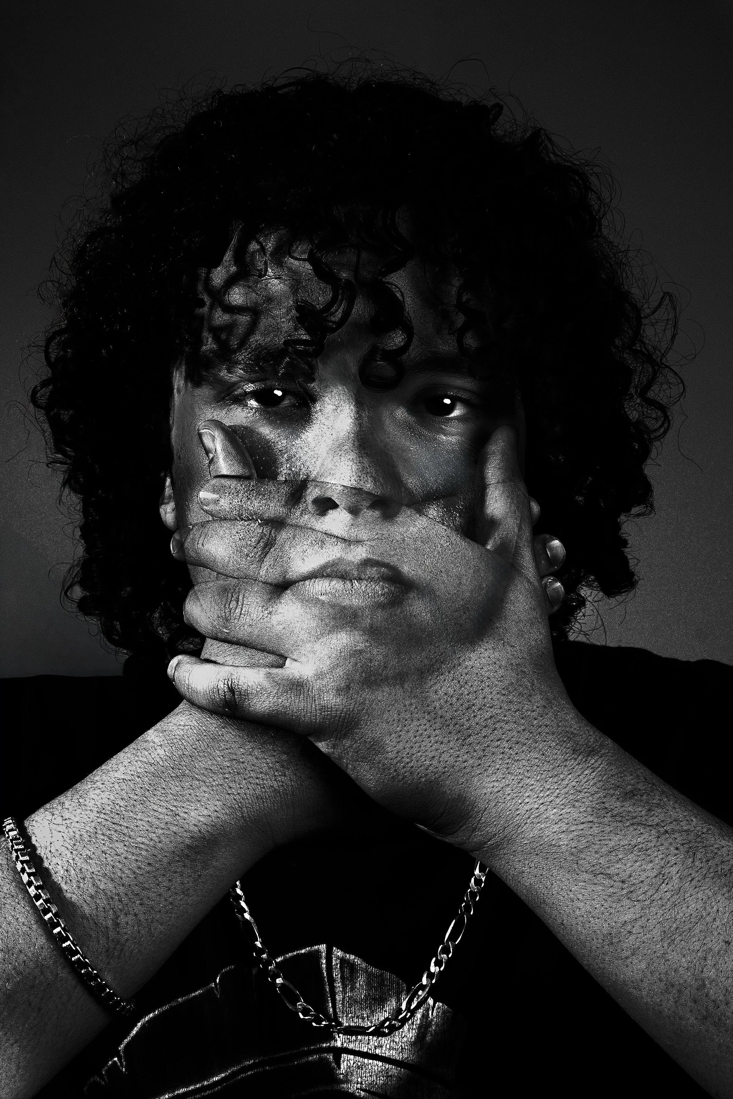 Black and white photo of a person with curly hair covering their mouth with both hands, wearing chains around their neck, and looking directly at the camera.