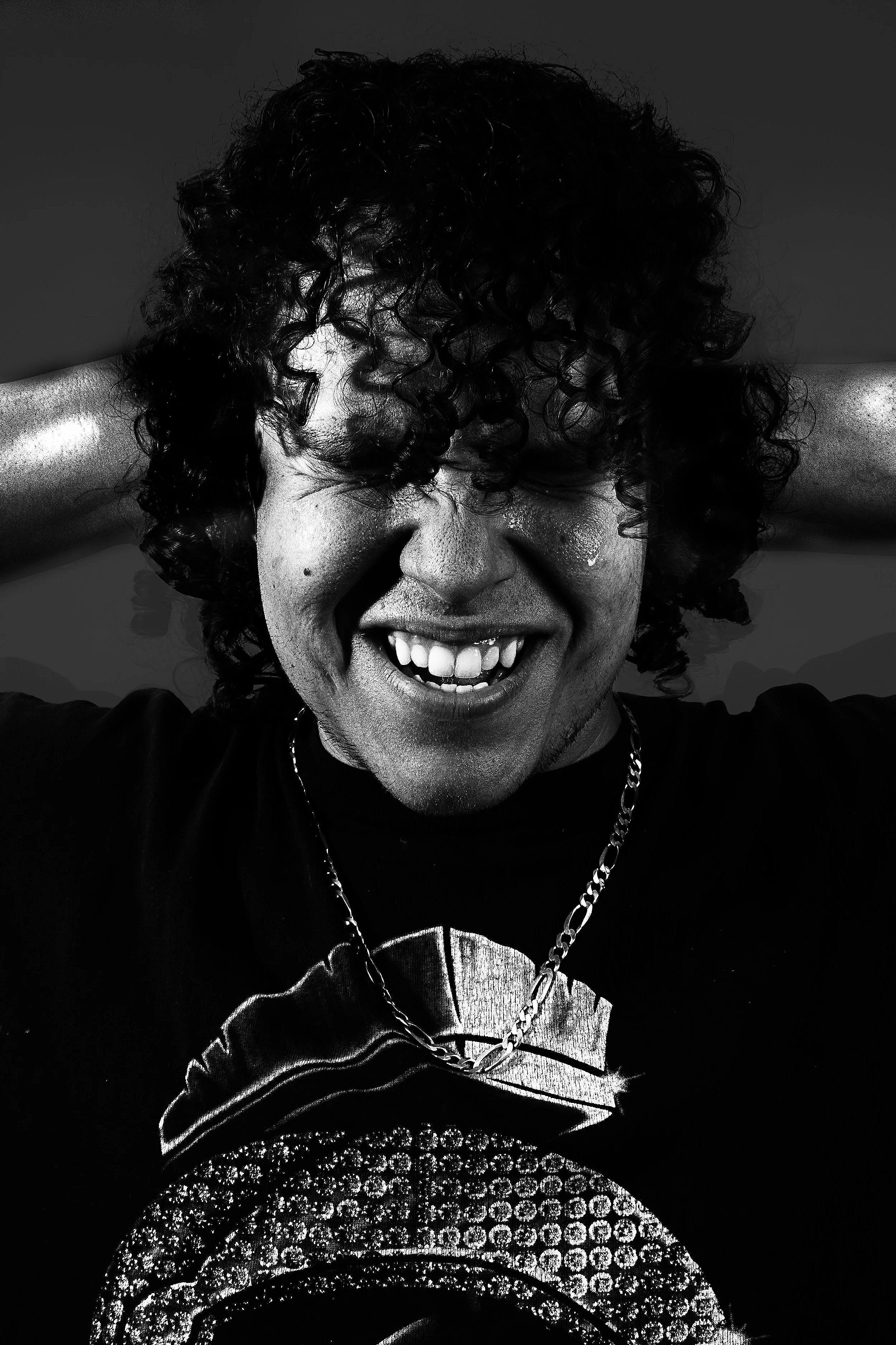 A person with curly hair and a big smile, wearing a black t-shirt with a graphic design, a chain necklace, and in a pose with their hands behind their head, in a black and white photo.