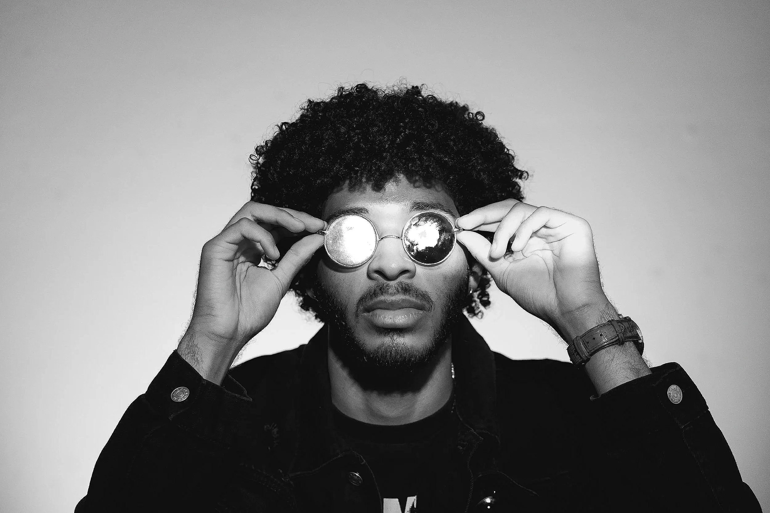 A black and white photo of a man with curly hair and a beard, wearing sunglasses and a dark denim jacket with metallic buttons, holding the sunglasses with both hands and looking directly at the camera.