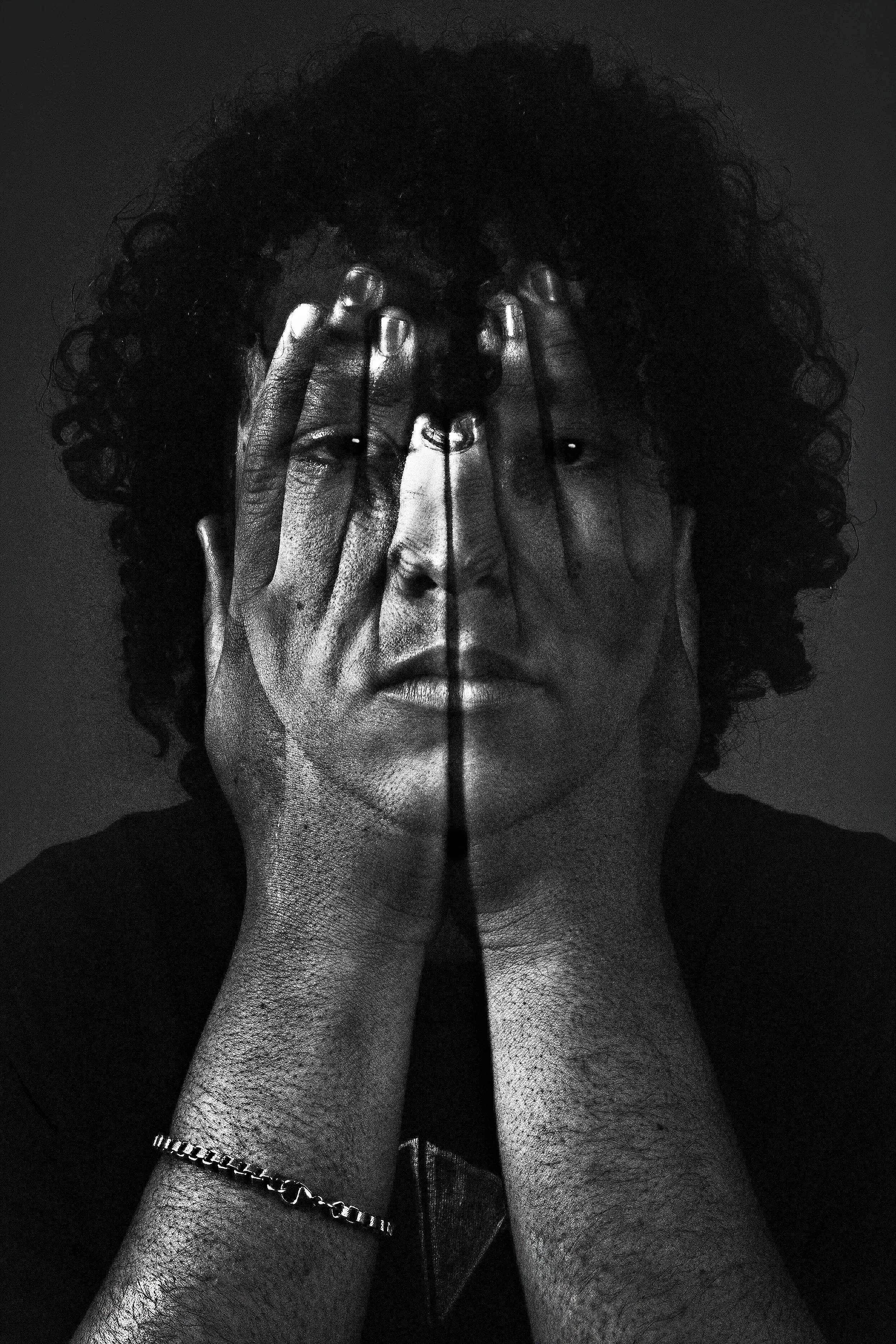 A black-and-white photograph of a person's face and hands covering their eyes. The individual has curly hair, bracelets, and a ring, with a dark background.