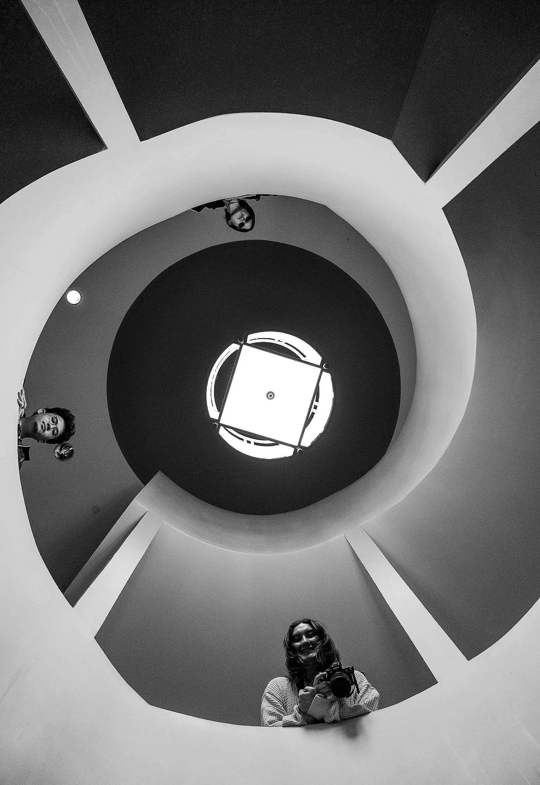 A woman taking a selfie with a camera in front of a mirror that reflects her and the staircase behind her in black and white.