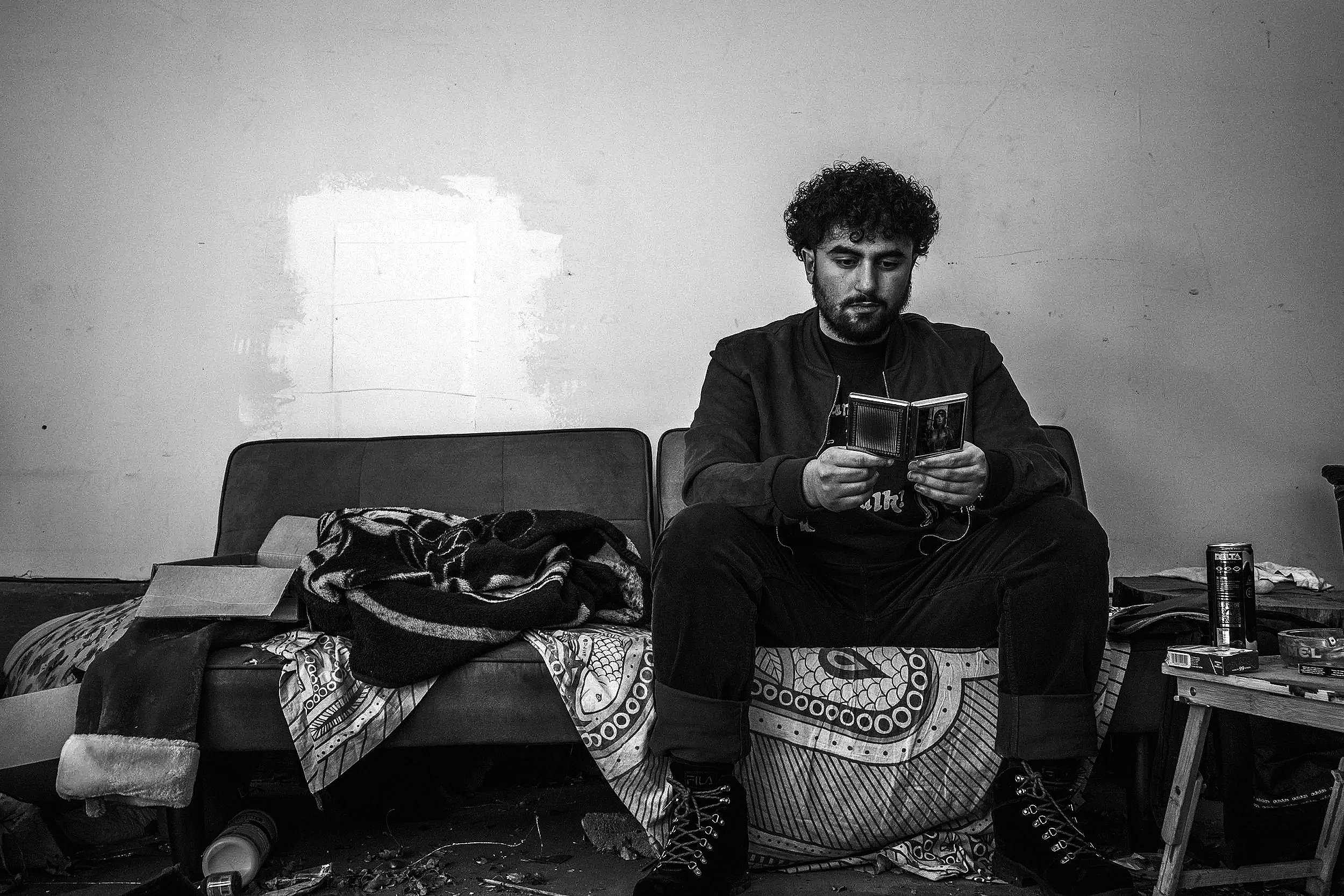 A man with curly hair and a beard sitting on a bed, reading a booklet or magazine. The room has a plain wall with some marks, and there are various items scattered around him, including a blanket, a can, and some books.