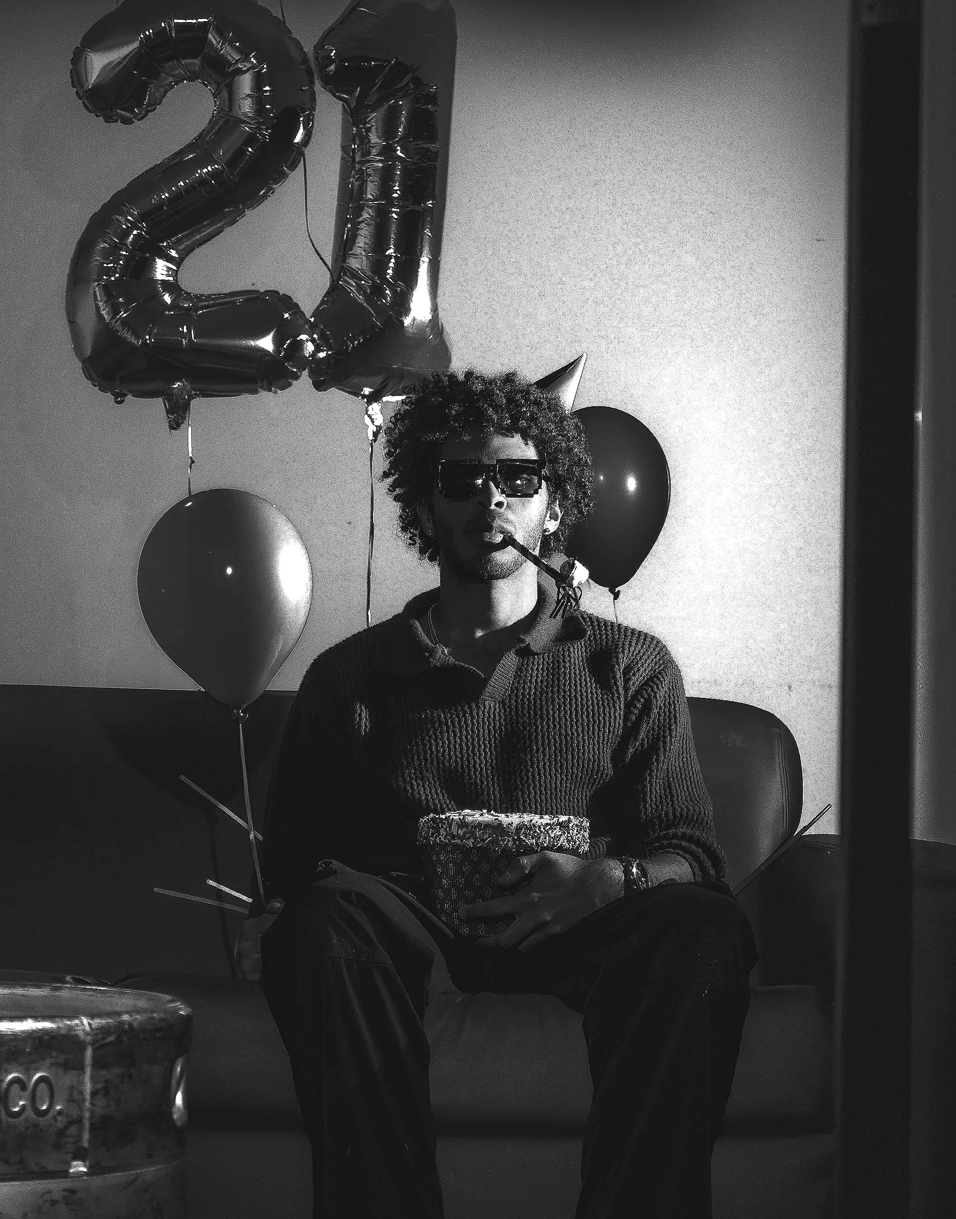 A person with curly hair wearing sunglasses and a party hat, sitting in front of a table with balloons and a large metallic number '20' balloon behind them.