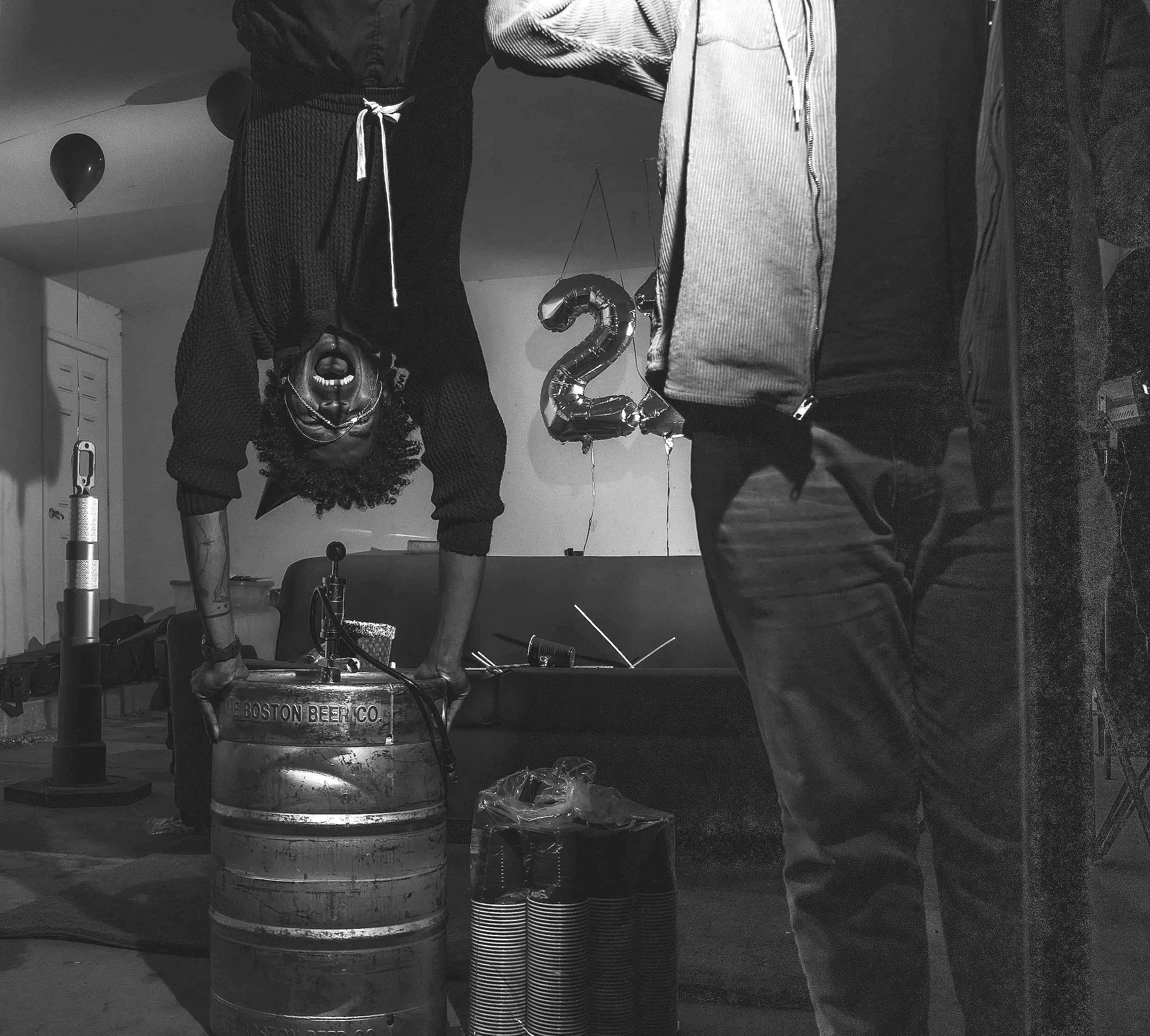 Two women leaning over a beer keg in a room decorated for a celebration, with balloons and a large number 2 balloon in the background.