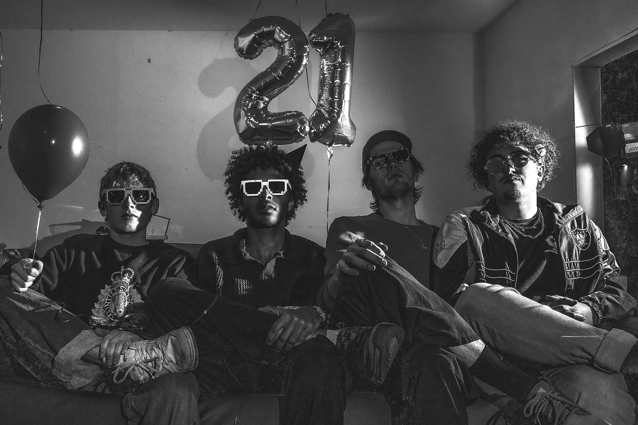 A group of four young men sitting together, dressed in casual and stylish clothing, wearing sunglasses with some holding balloons, and there are large balloon numbers '2' and '1' floating above them, suggesting a celebration.