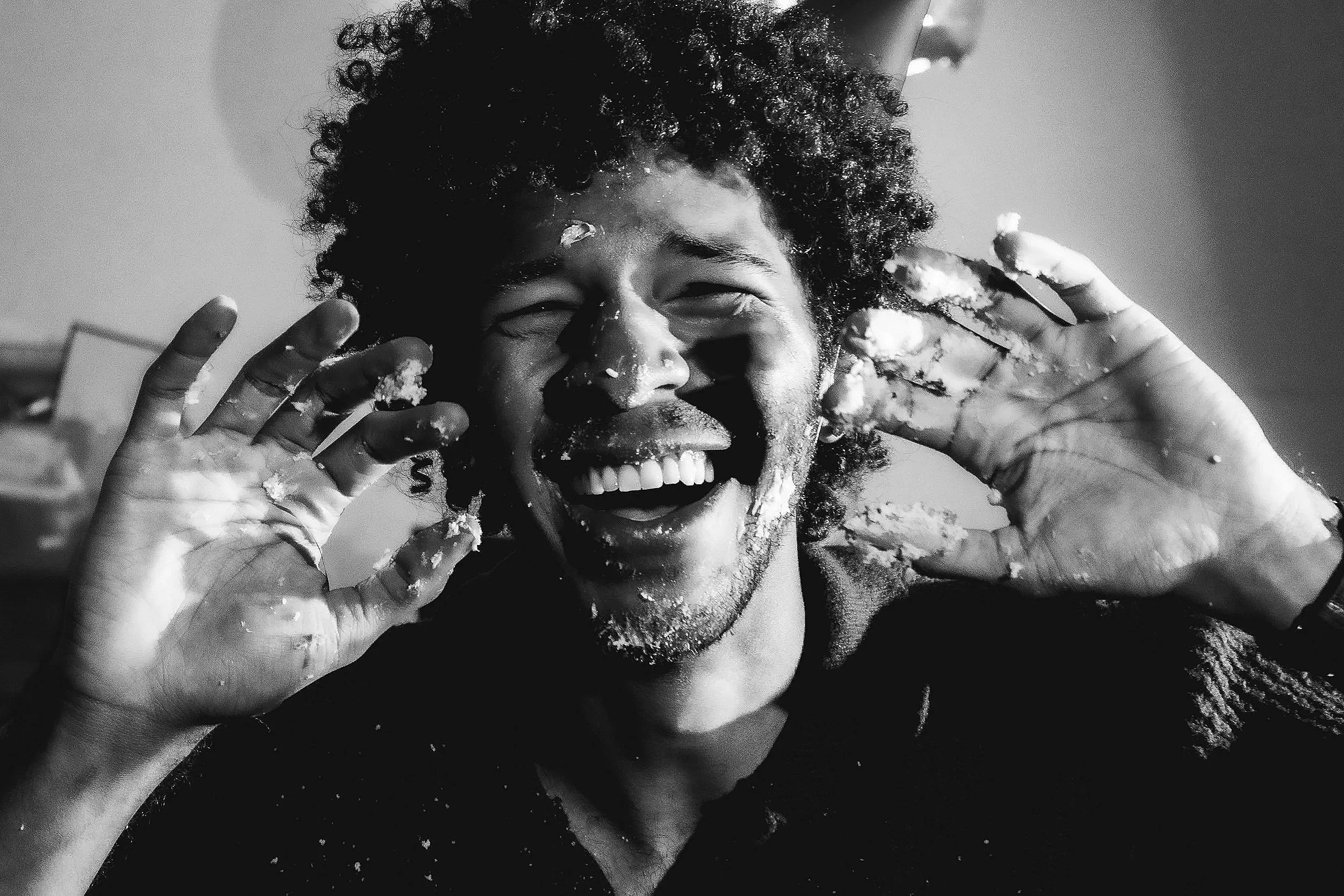 A person with curly hair and a wide smile, covered in cake or frosting, with hands near their face.