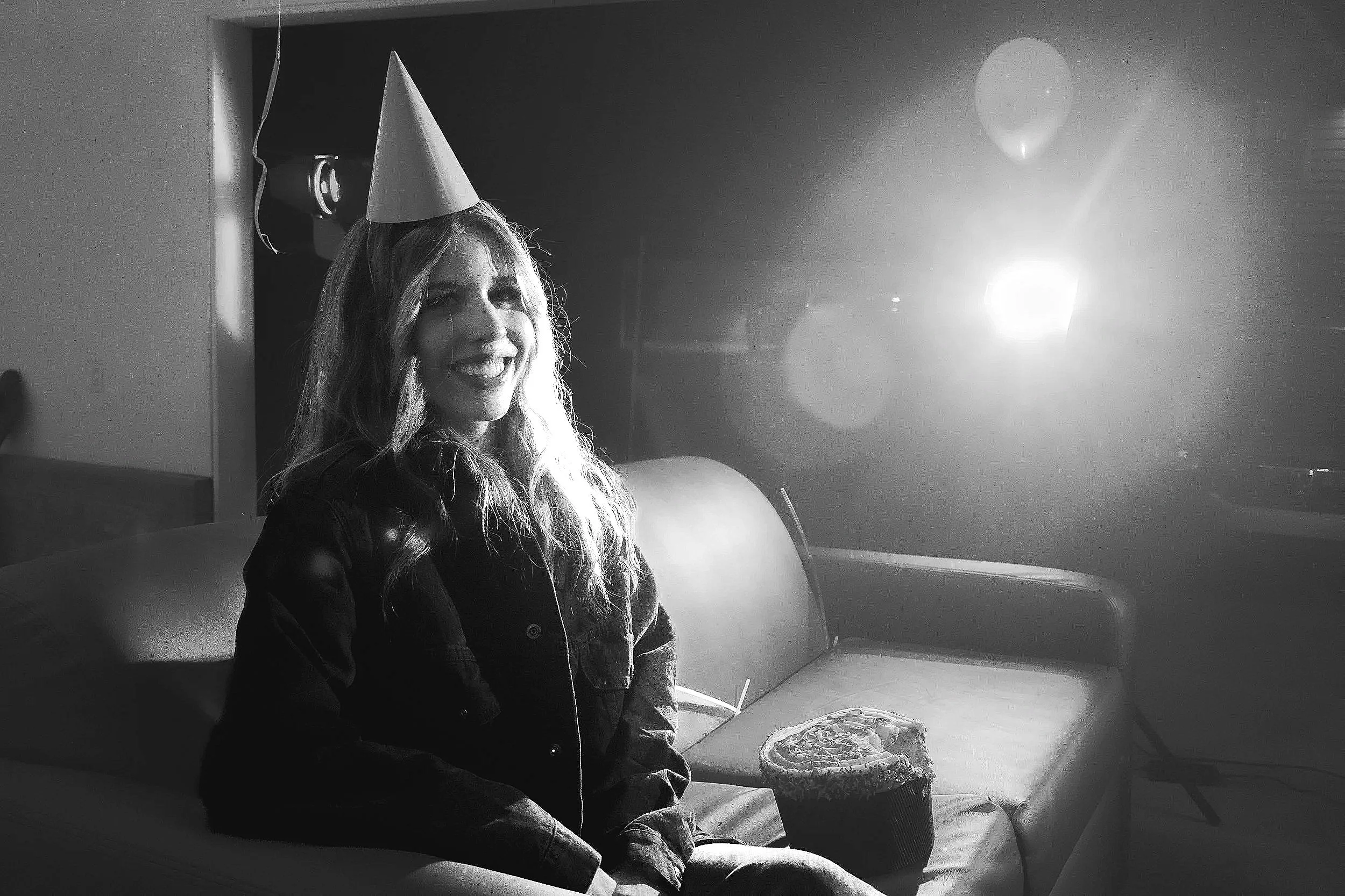 A young woman with long, wavy hair smiling while wearing a party hat, sitting on a couch with a birthday cake on her lap. The photo is in black and white with a bright light source behind her.