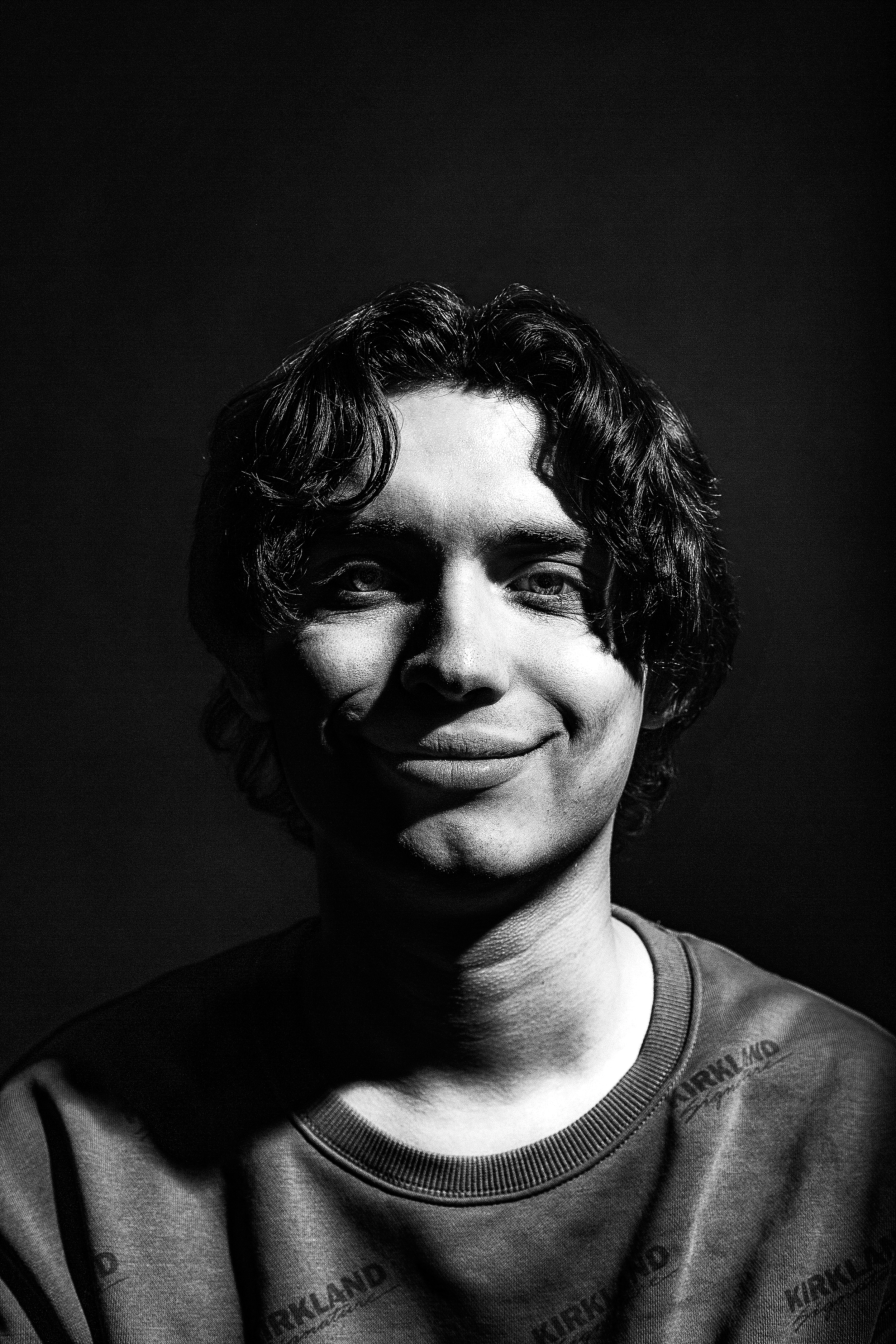 A black and white photo of a young person with short, wavy hair, smiling and wearing a shirt with text on it.