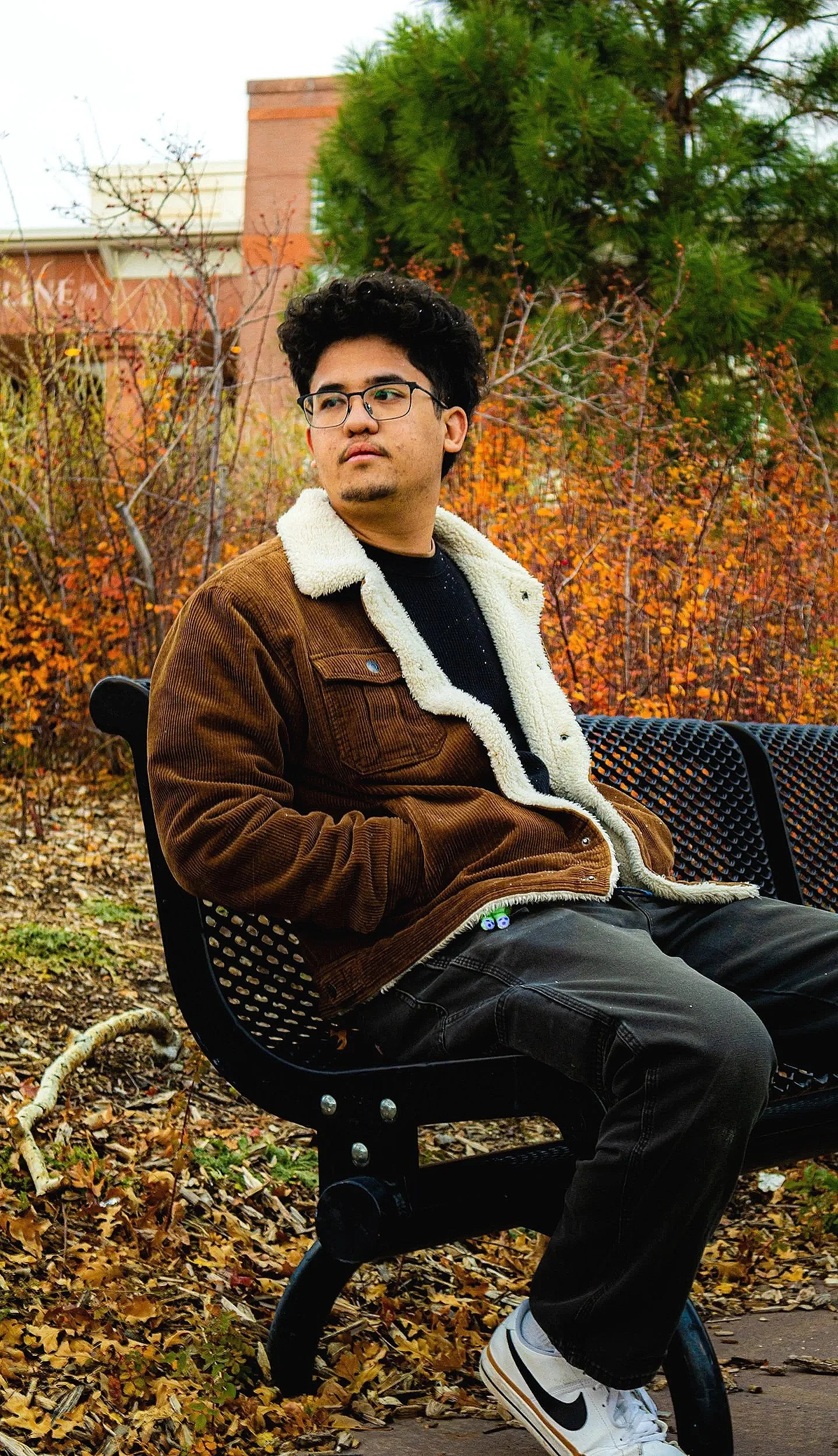 A young man with dark curly hair, glasses, and a short beard sits on a black metal park bench covered in fallen autumn leaves. He is wearing a brown corduroy jacket with a white shearling collar, a black shirt, black jeans, and white Nike sneakers. T