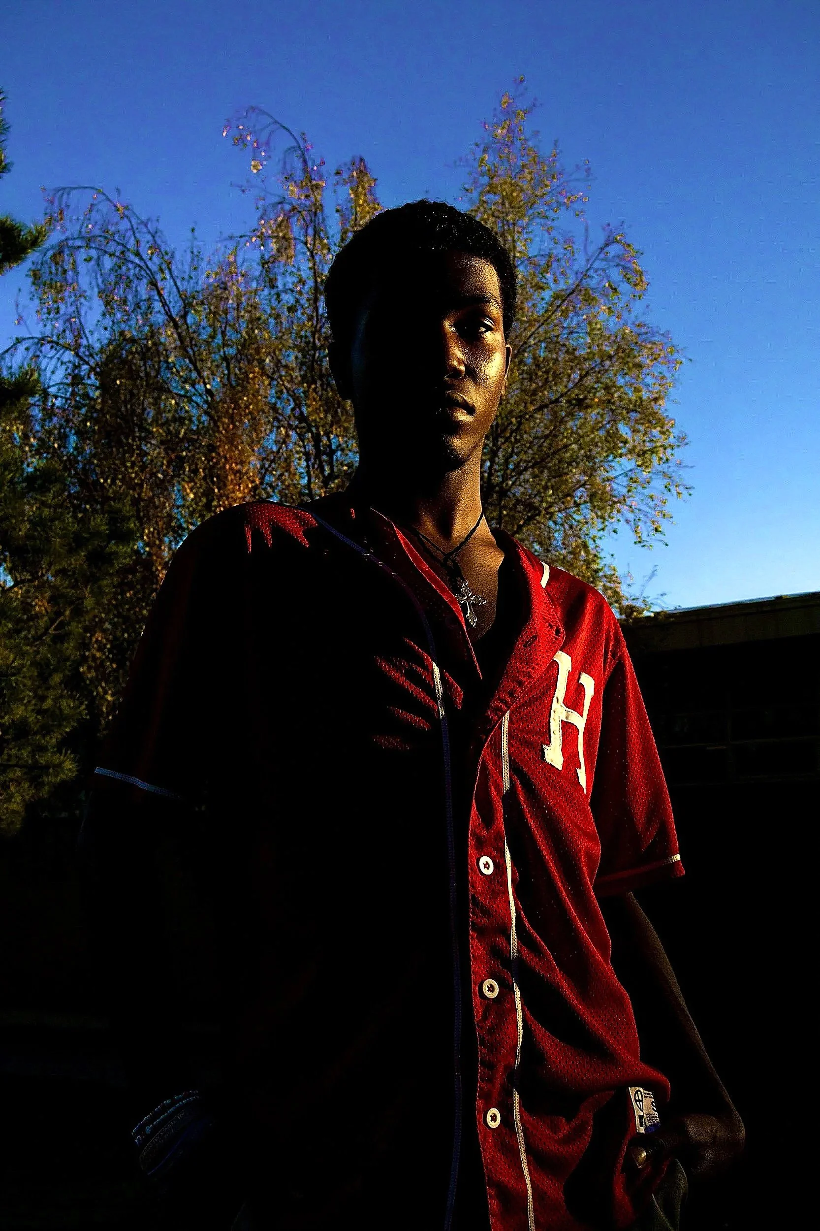 Young person with dark skin and short curly hair standing outdoors during sunset, wearing a red sports jacket with white lettering and a black necklace, with trees and a clear blue sky in the background.