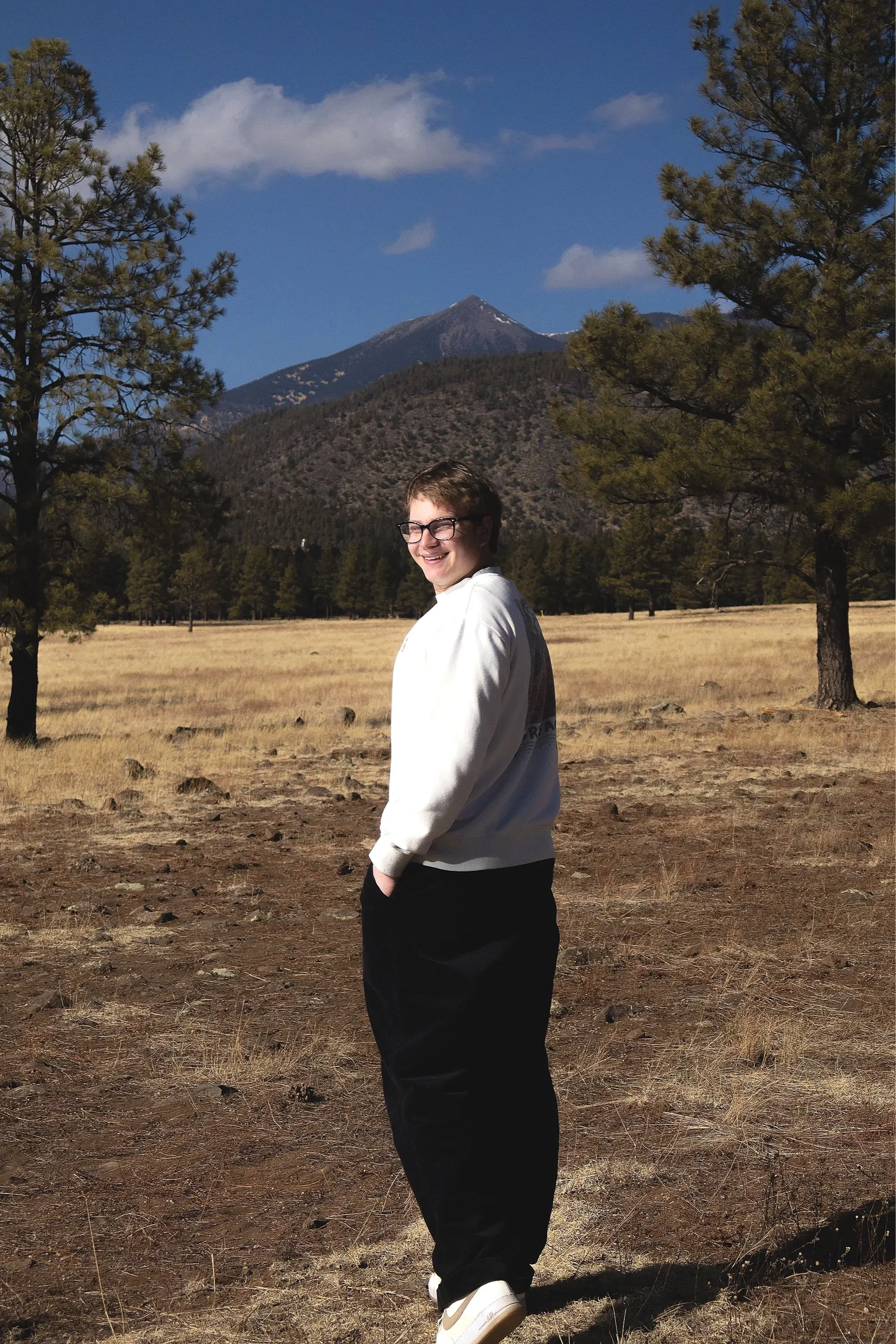 Young man with glasses smiling, standing outdoors in a natural landscape with trees and mountains.