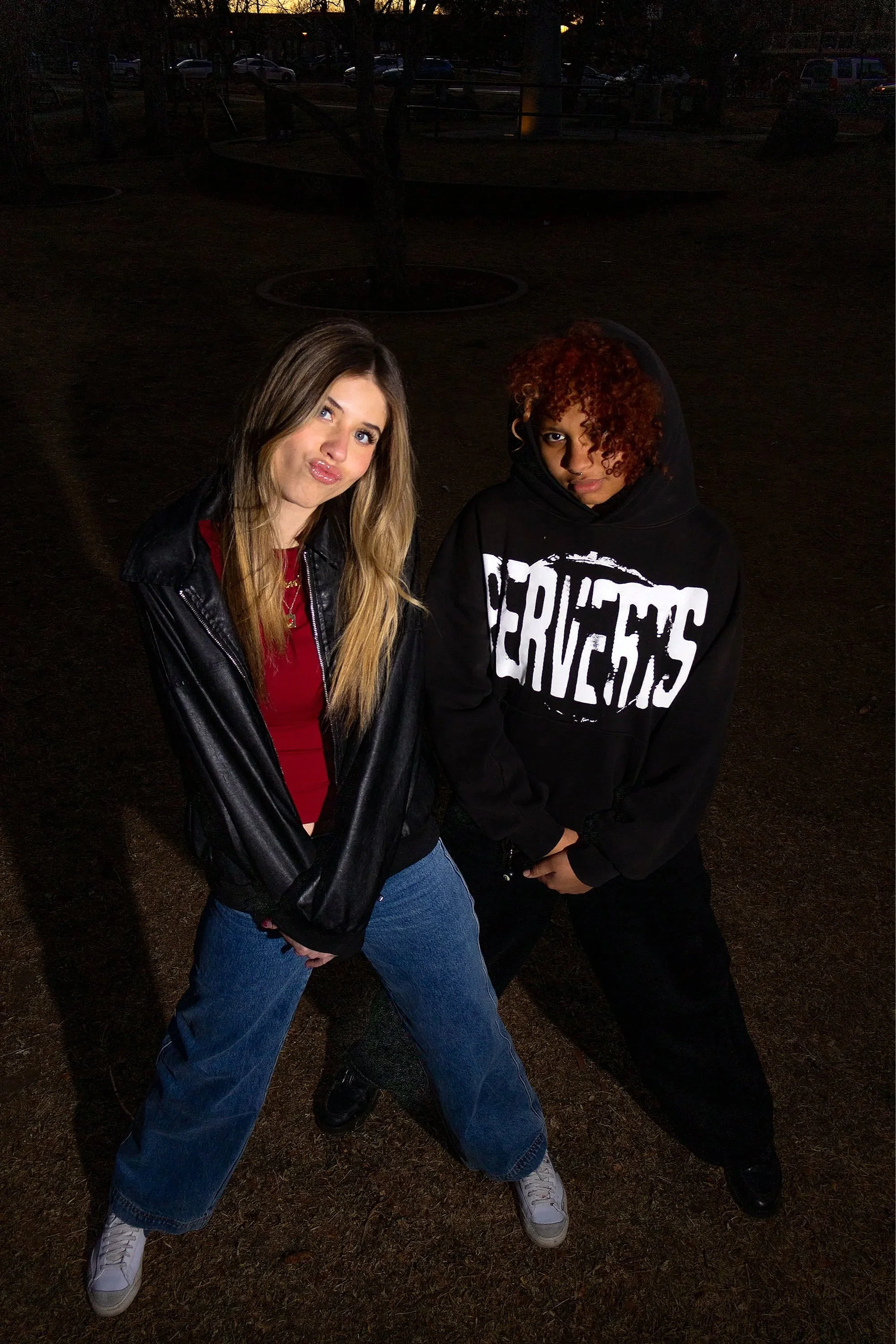 Two young women posing outdoors at dusk, one with long blond hair in a leather jacket and red top, the other with curly red hair in a black hoodie with white text, standing with their legs apart and their hands near their hips.