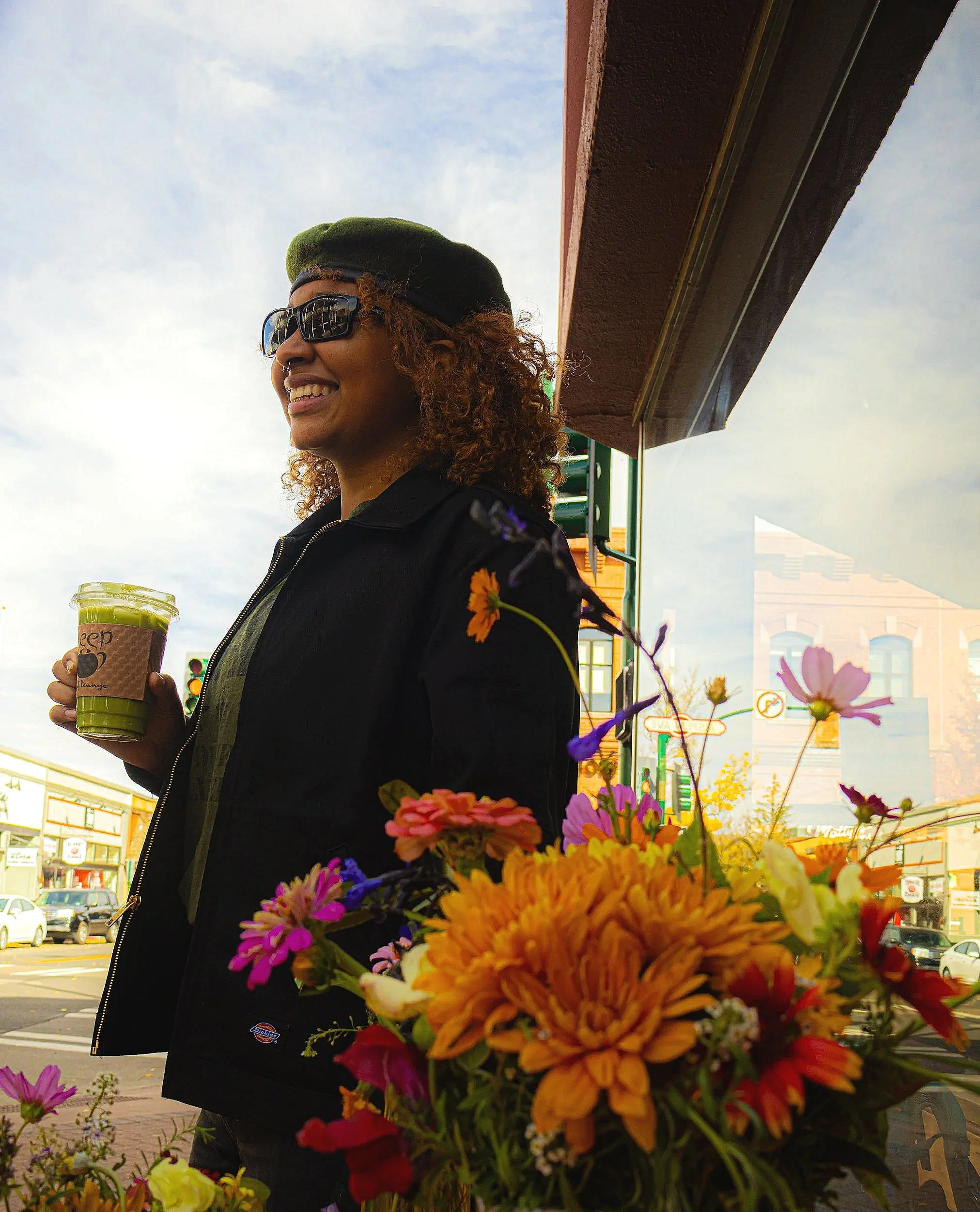 A smiling woman with curly hair, wearing sunglasses and a black beret, holds a green smoothie in a disposable cup with a brown sleeve. She stands outside a shop next to a flower display. The background shows a street scene with buildings, cars, and t
