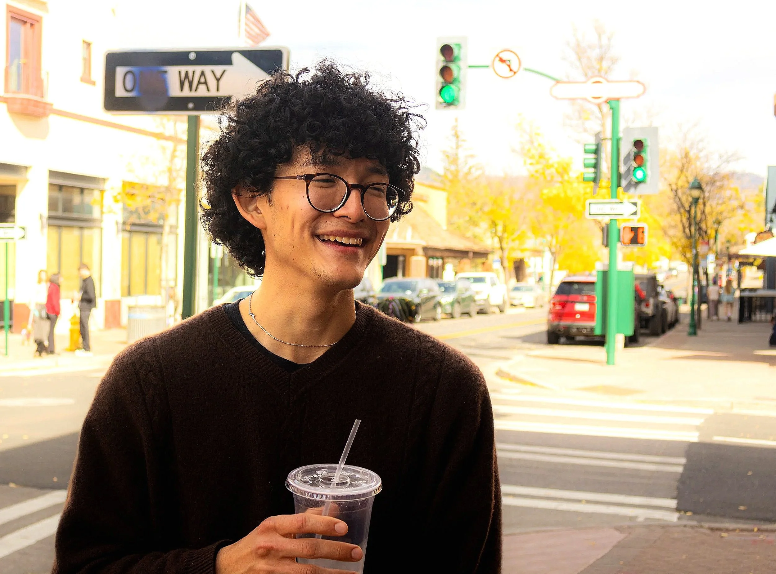 A young man with curly black hair and glasses smiling while holding a plastic cup with a drink and a straw on a city street with traffic lights and buildings in the background.