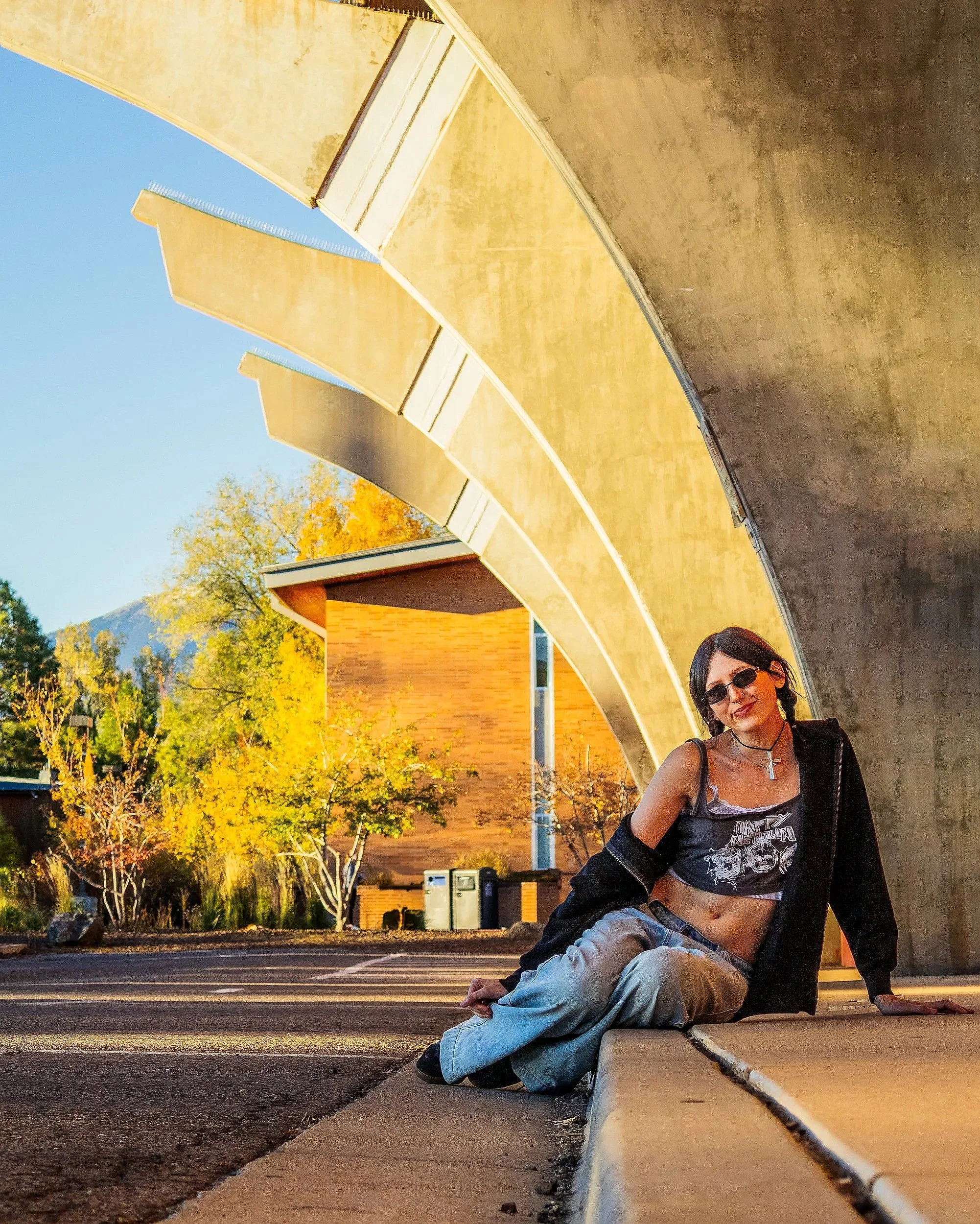 A woman with dark hair, wearing sunglasses, a black jacket, and a graphic tank top, sitting on the curb with her friend lying down in front of her. They are outdoors under a yellow modern architectural structure, with trees and a building in the back