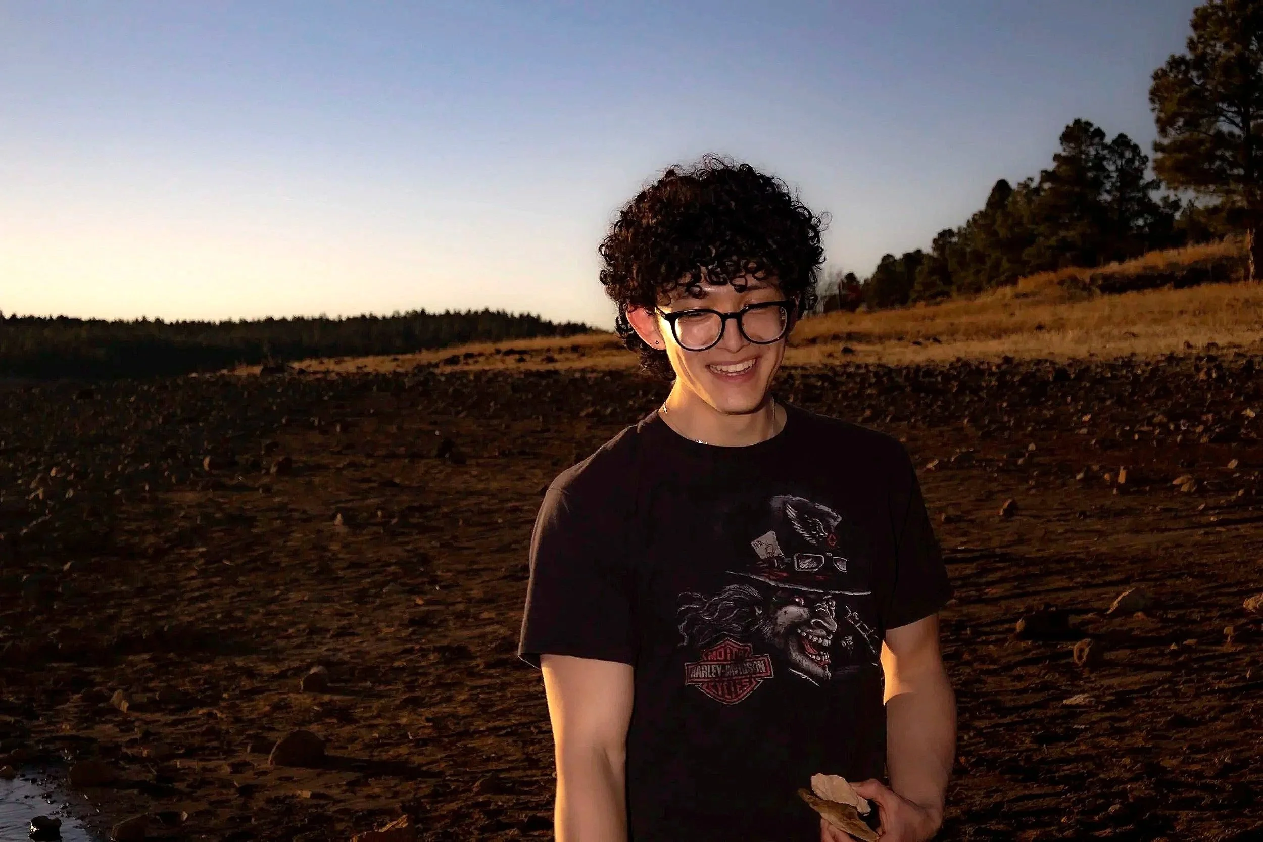 A smiling young person with curly dark hair and glasses stands outdoors during sunset, with trees and a rocky landscape in the background.