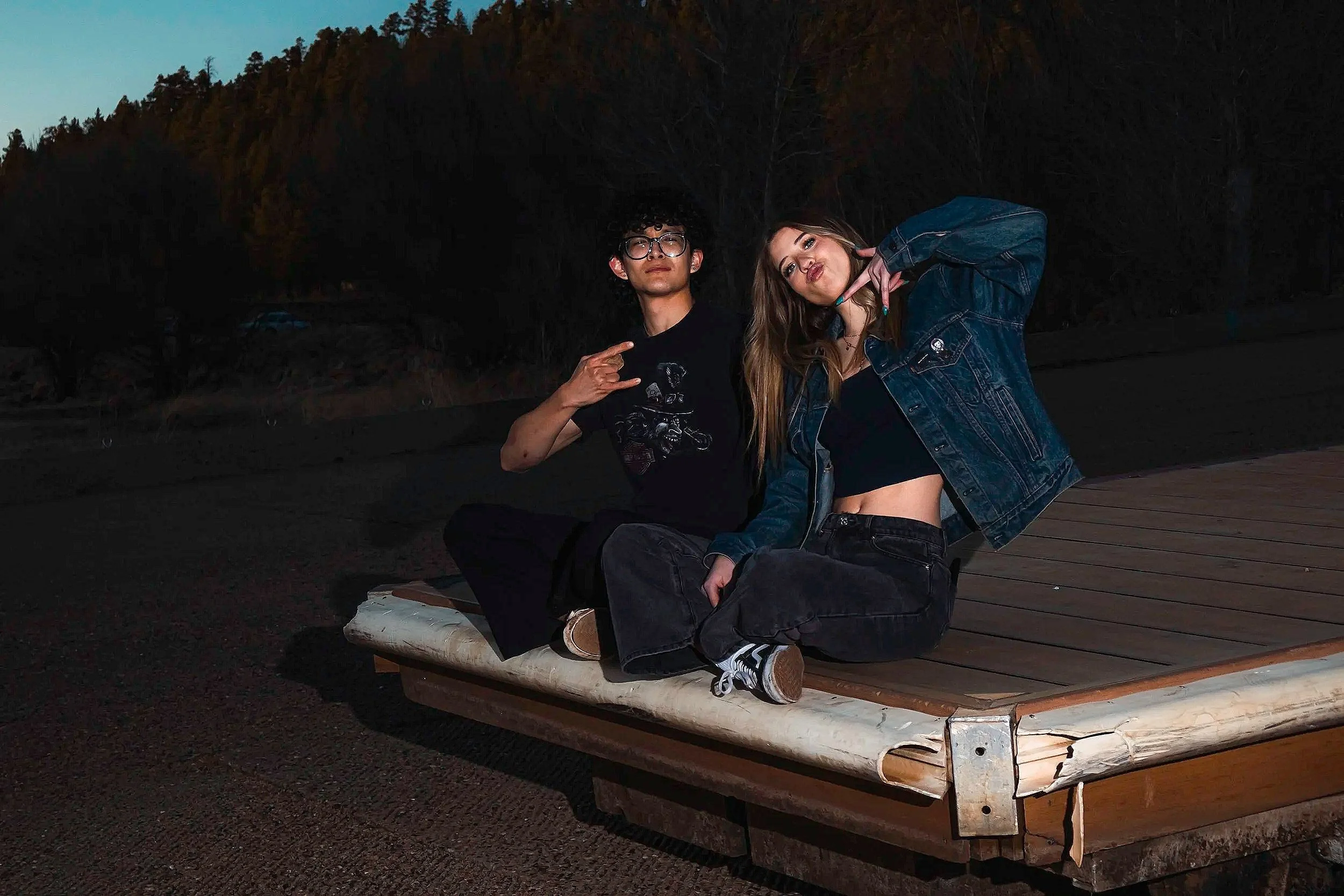 Two young people sitting outdoors on a wooden platform at night, with trees in the background. The person on the left has curly hair, glasses, and is making a hand gesture, while the person on the right has long hair and is making a peace sign near t