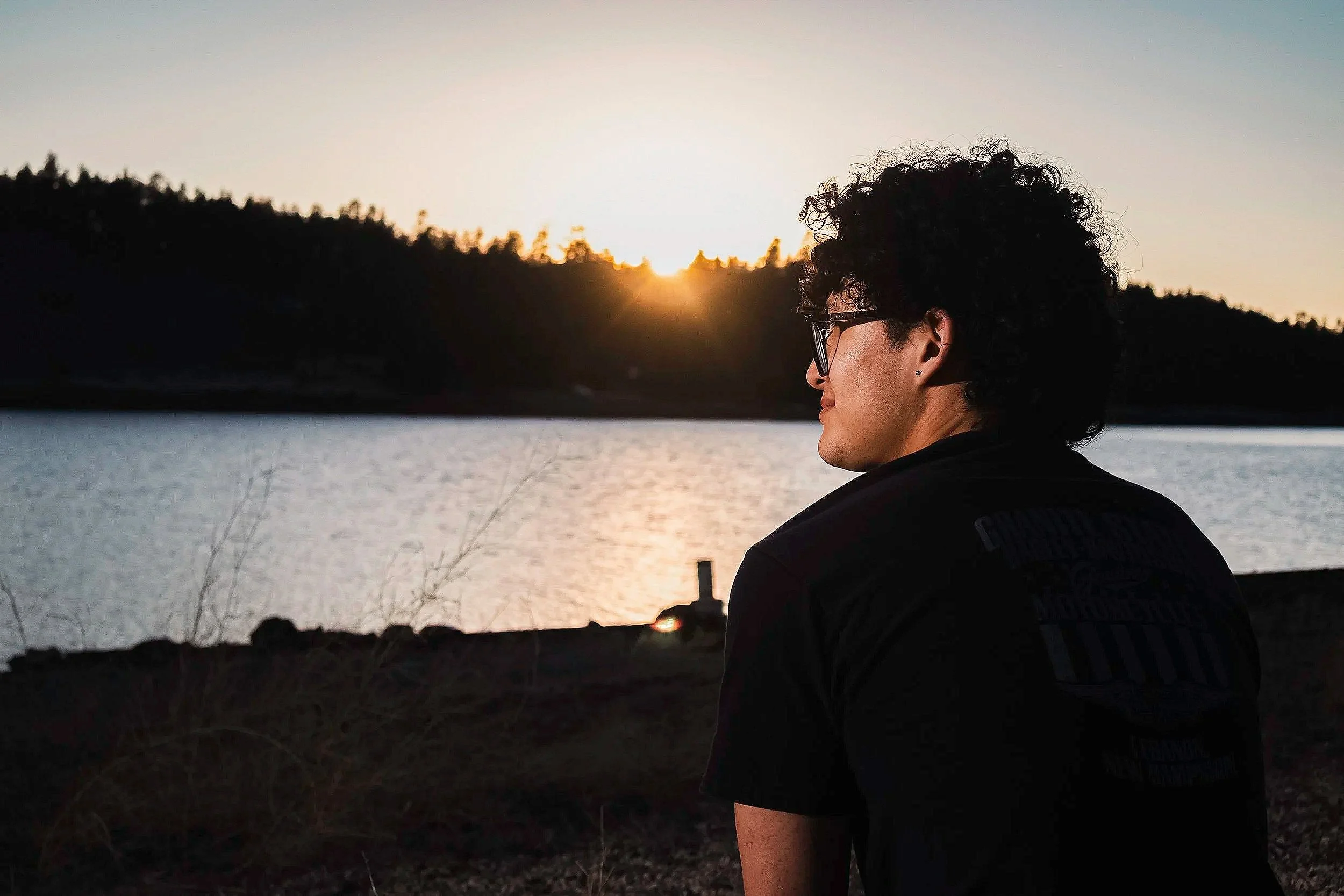 A person with curly hair and glasses sitting outdoors by a body of water during sunset, with a silhouette of trees in the background.