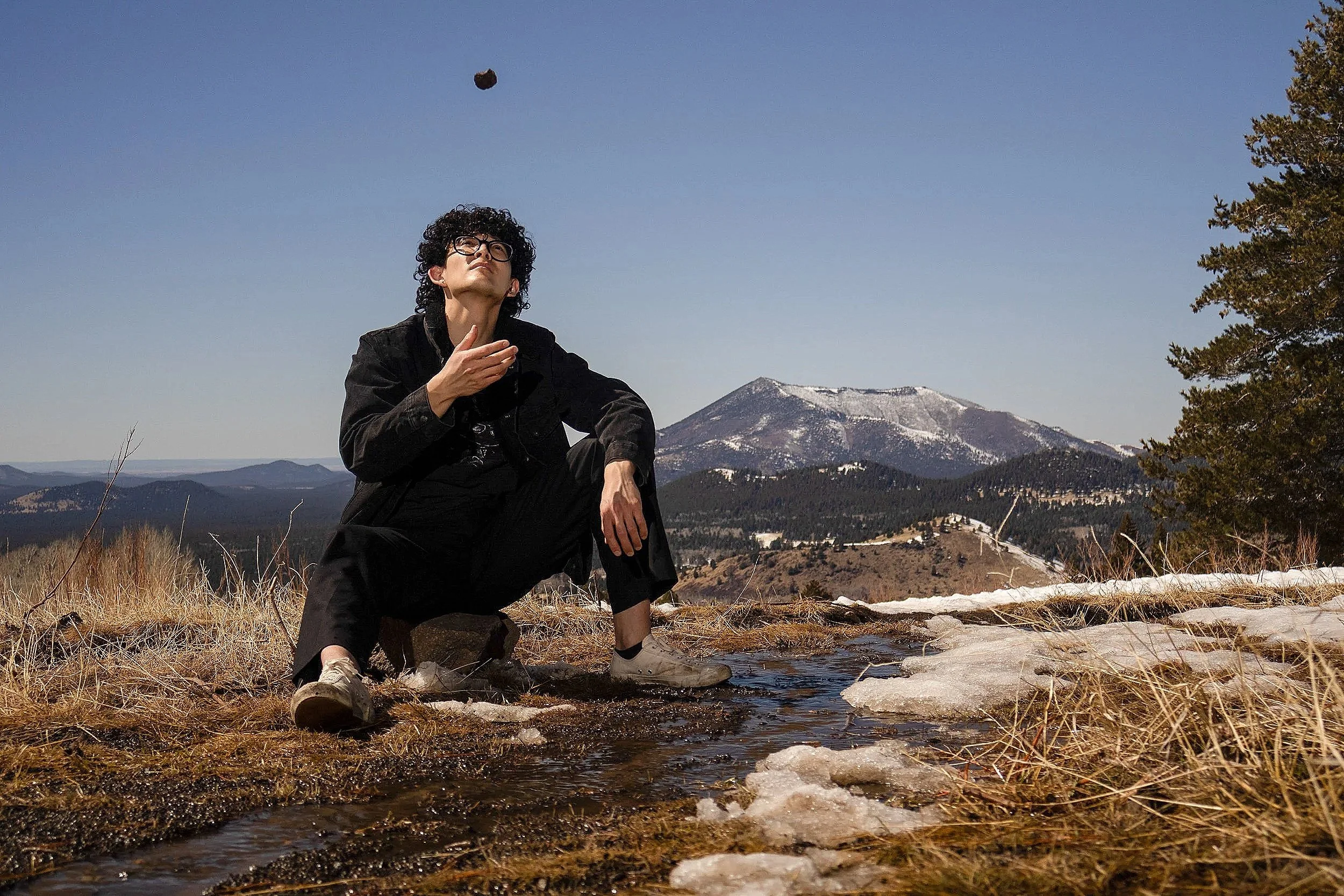 A person with curly hair and glasses sitting on a rock next to a small creek in a mountainous landscape with snow-capped peaks in the background.