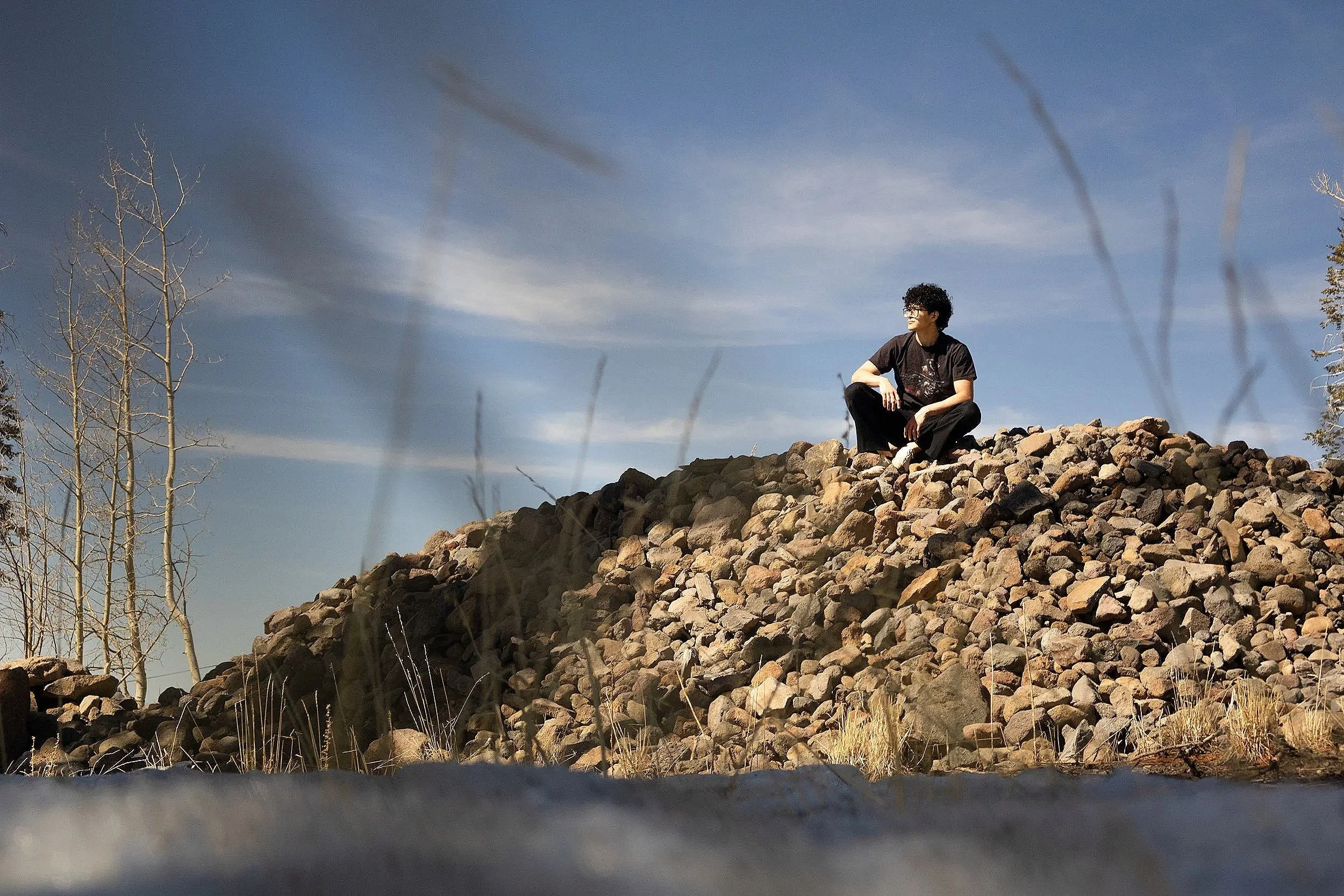 A person with curly black hair wearing black clothing and glasses sits on a pile of rocks outdoors, with a clear blue sky and sparse trees in the background.