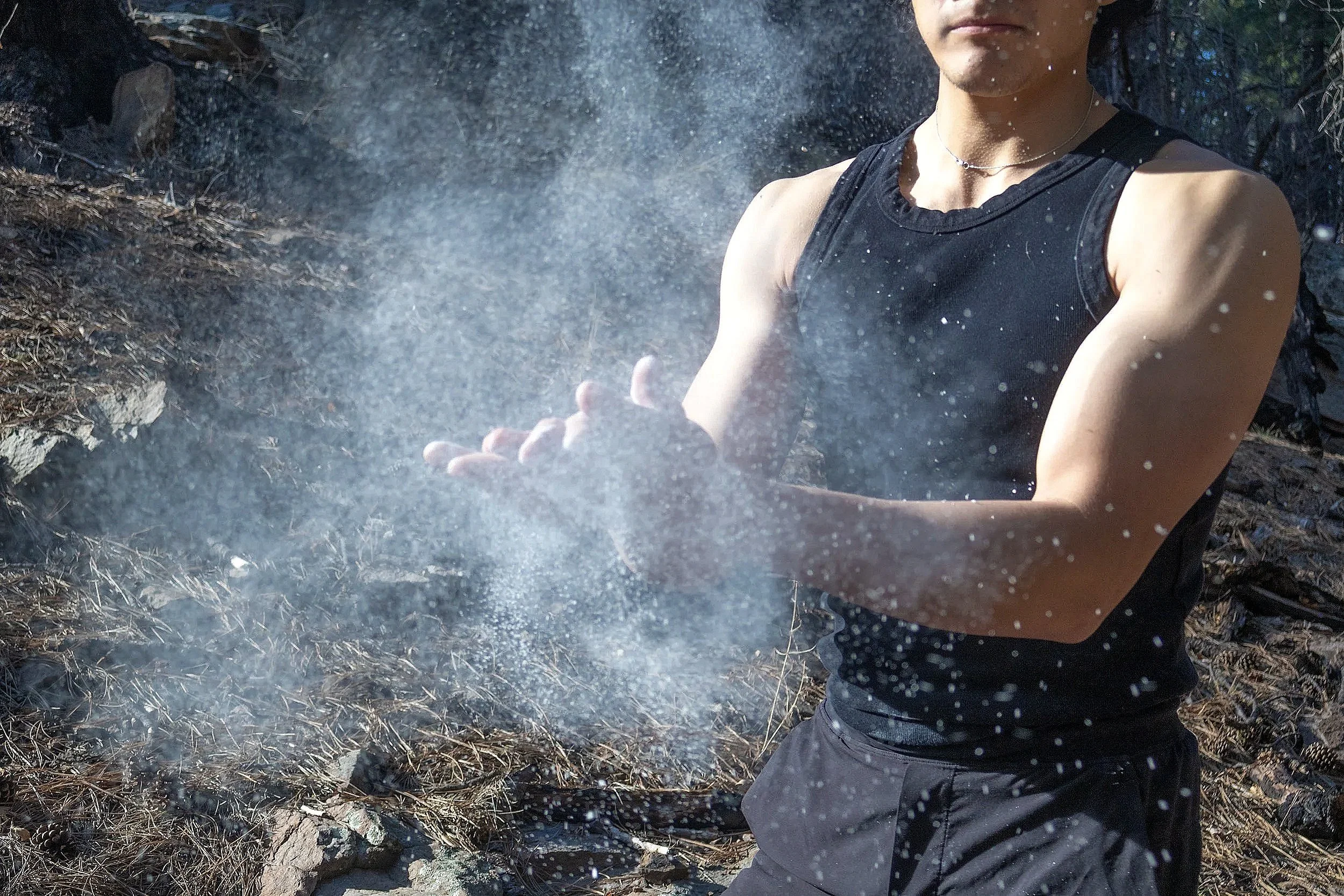 A person wearing a black sleeveless shirt and dark pants, standing outdoors amidst charred ground and ashes, with dust particles in the air.