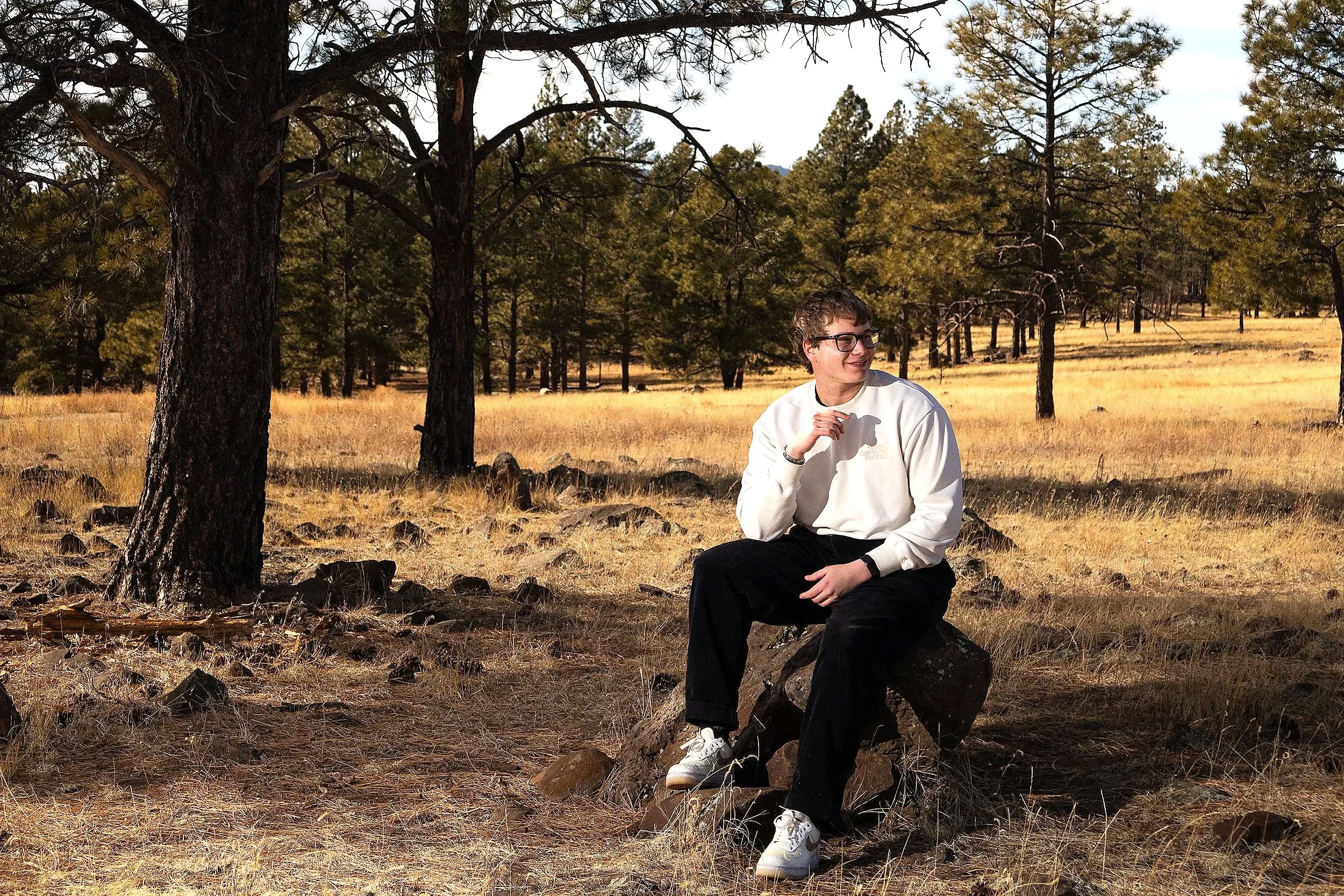 A young man with glasses and a white sweatshirt sitting on a rock in a forest with tall trees and golden grass.