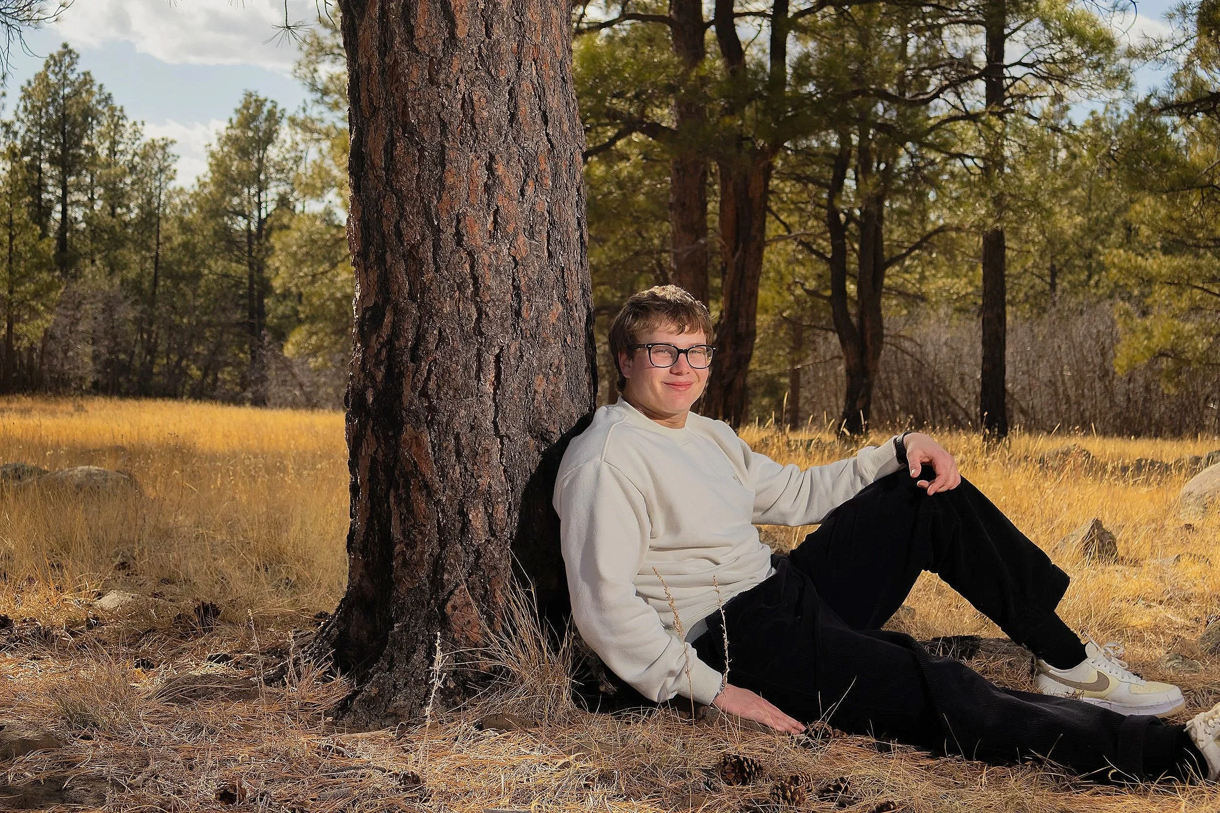 A young man with glasses, wearing a white sweatshirt and black pants, sitting on the ground in a forest, leaning against a large tree trunk, with a relaxed expression and smile, surrounded by tall trees and golden grass.