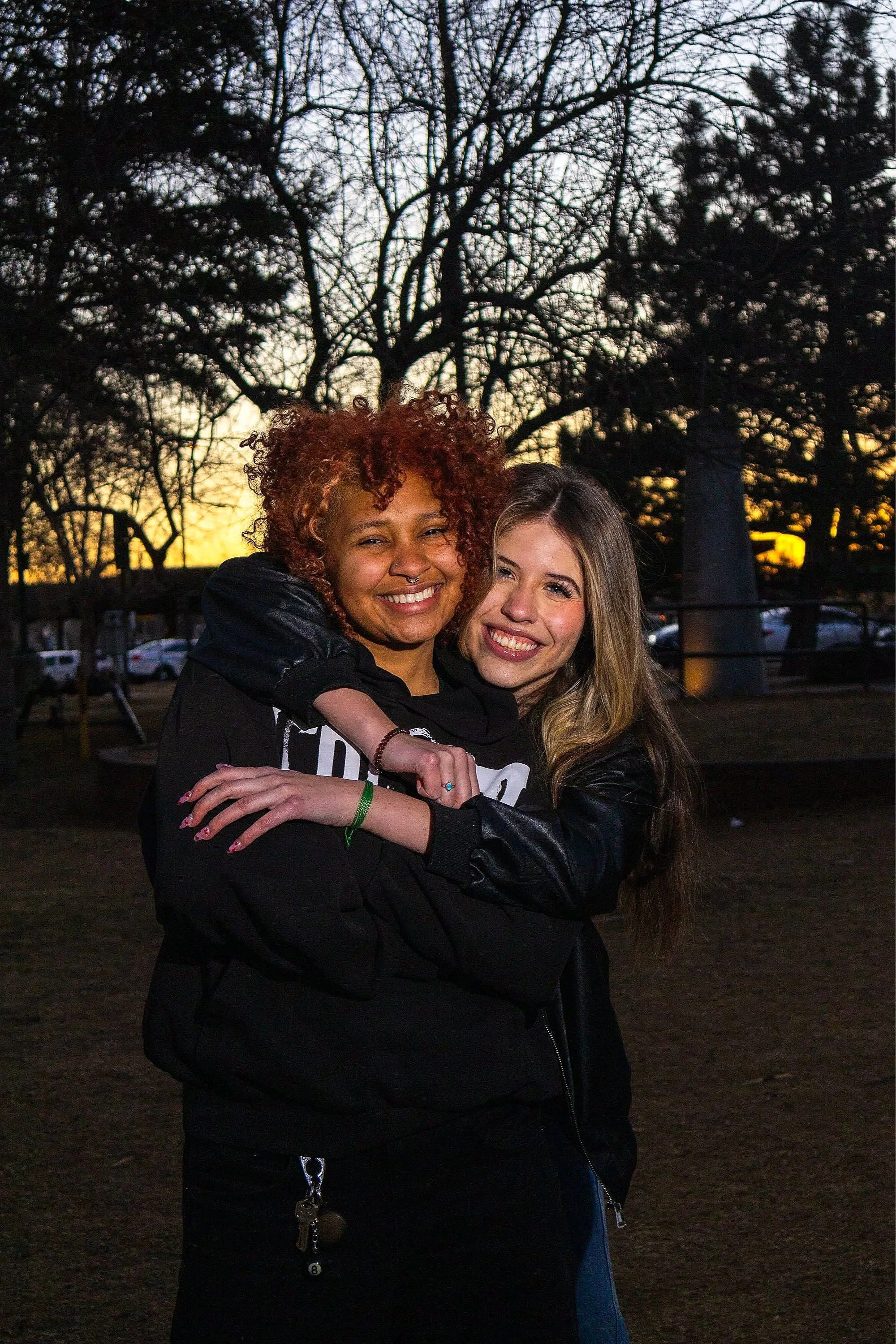 Two smiling women hugging outdoors during sunset, with leafless trees in the background.