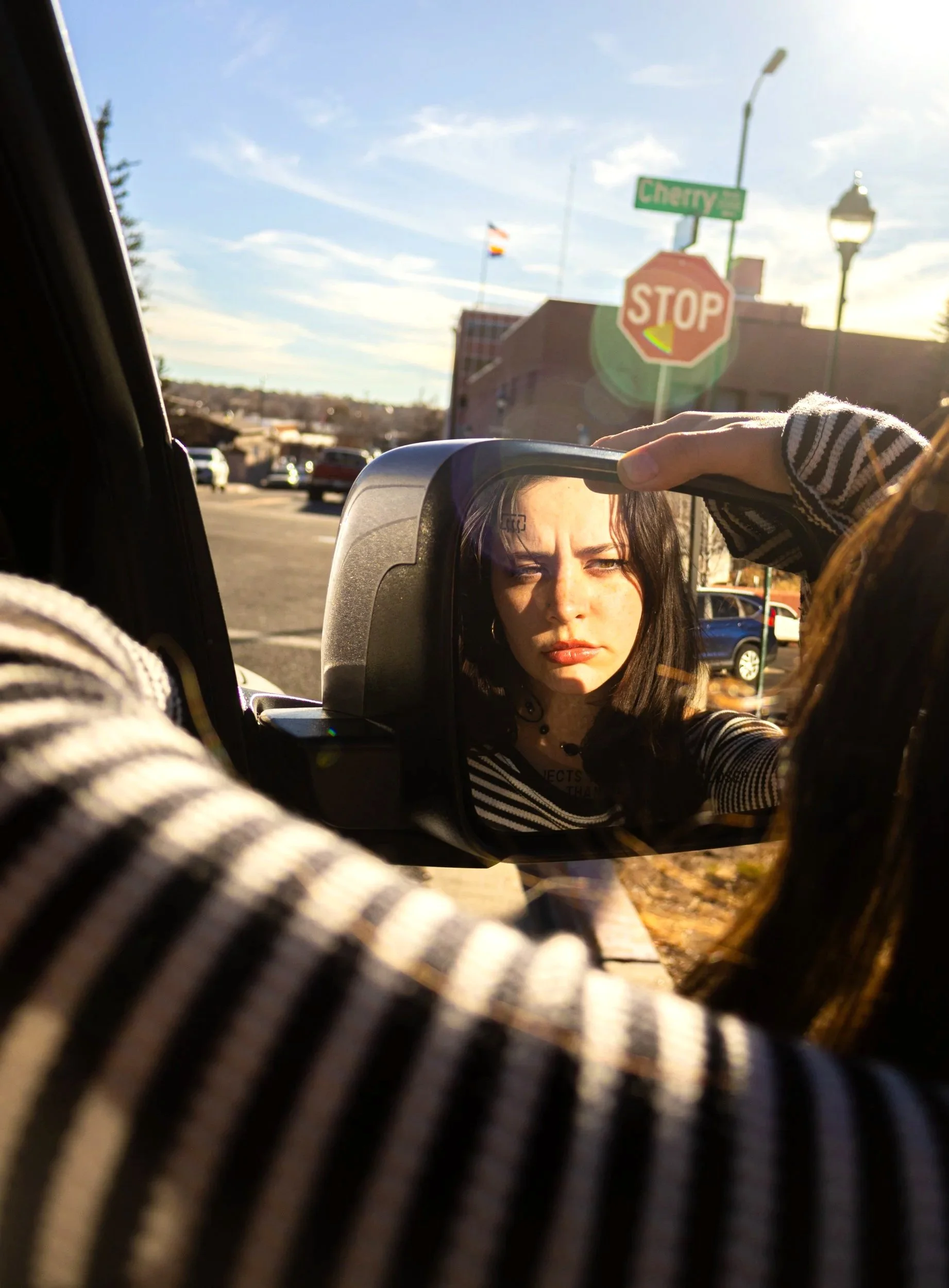 A woman is looking into a side view mirror of a vehicle, with a street, stop sign, and street signs visible in the background, during sunset.