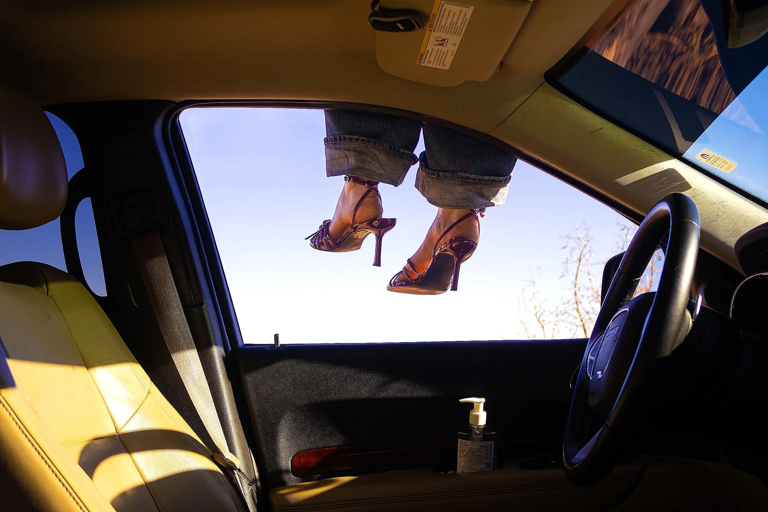 The interior of a car viewed from the back seat, with a pair of legs wearing high heels extending out of the sunroof.