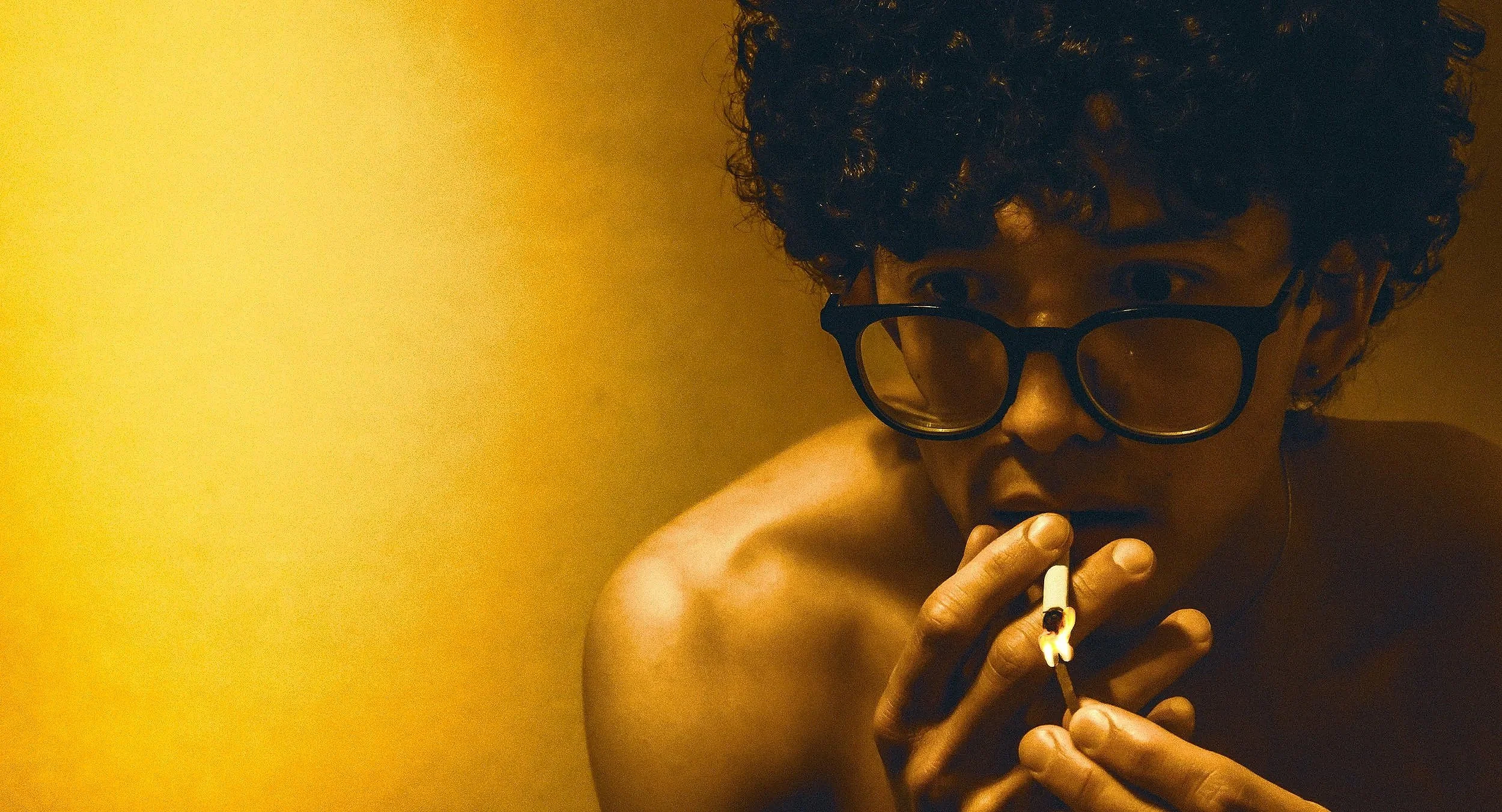 A woman with curly hair and glasses smokes a cigarette against a yellow background.