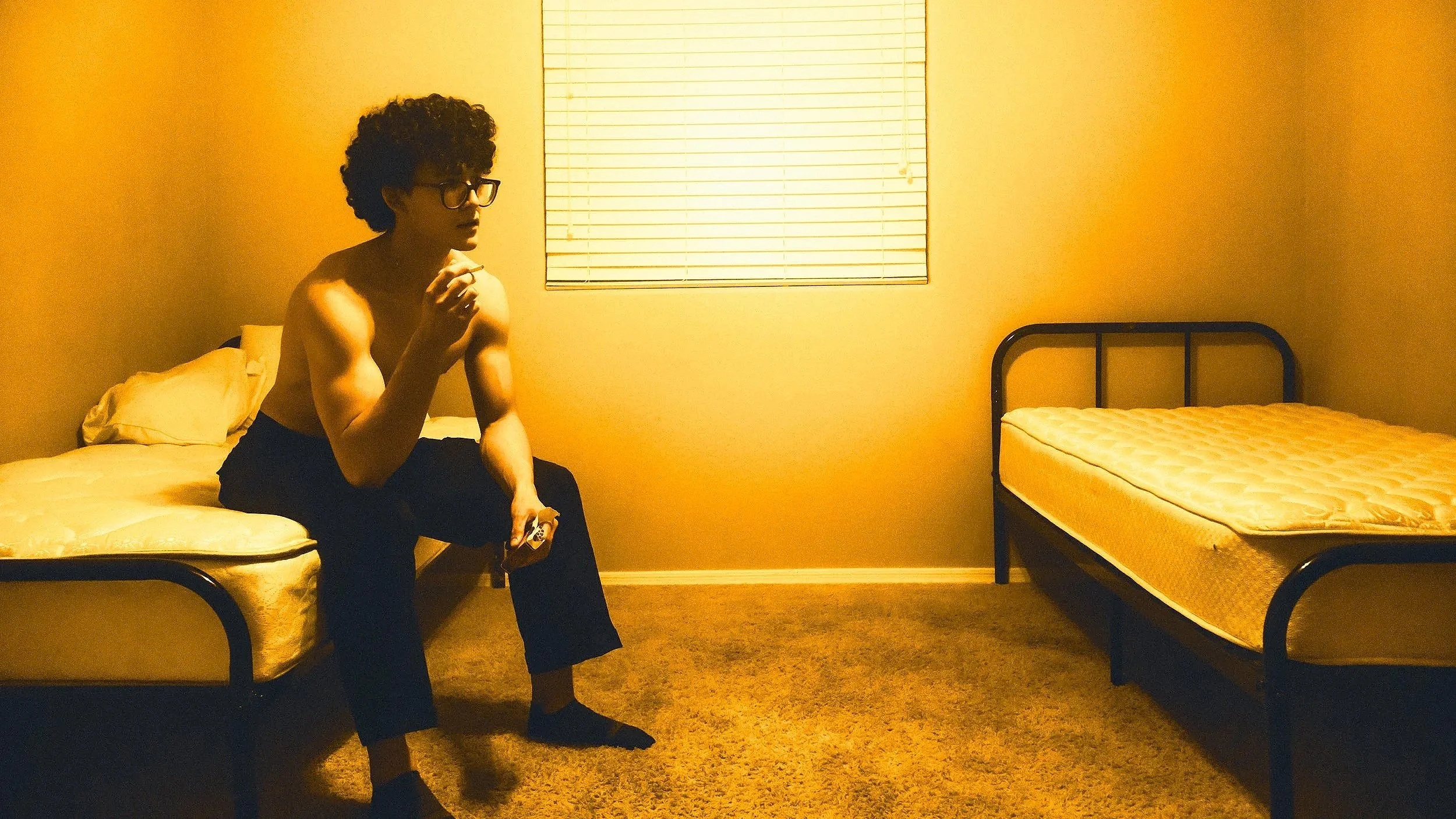 A young man with curly hair and glasses sitting on a bed in a sparsely furnished room with two metal-framed beds and a window with closed blinds, illuminated by warm yellow light.