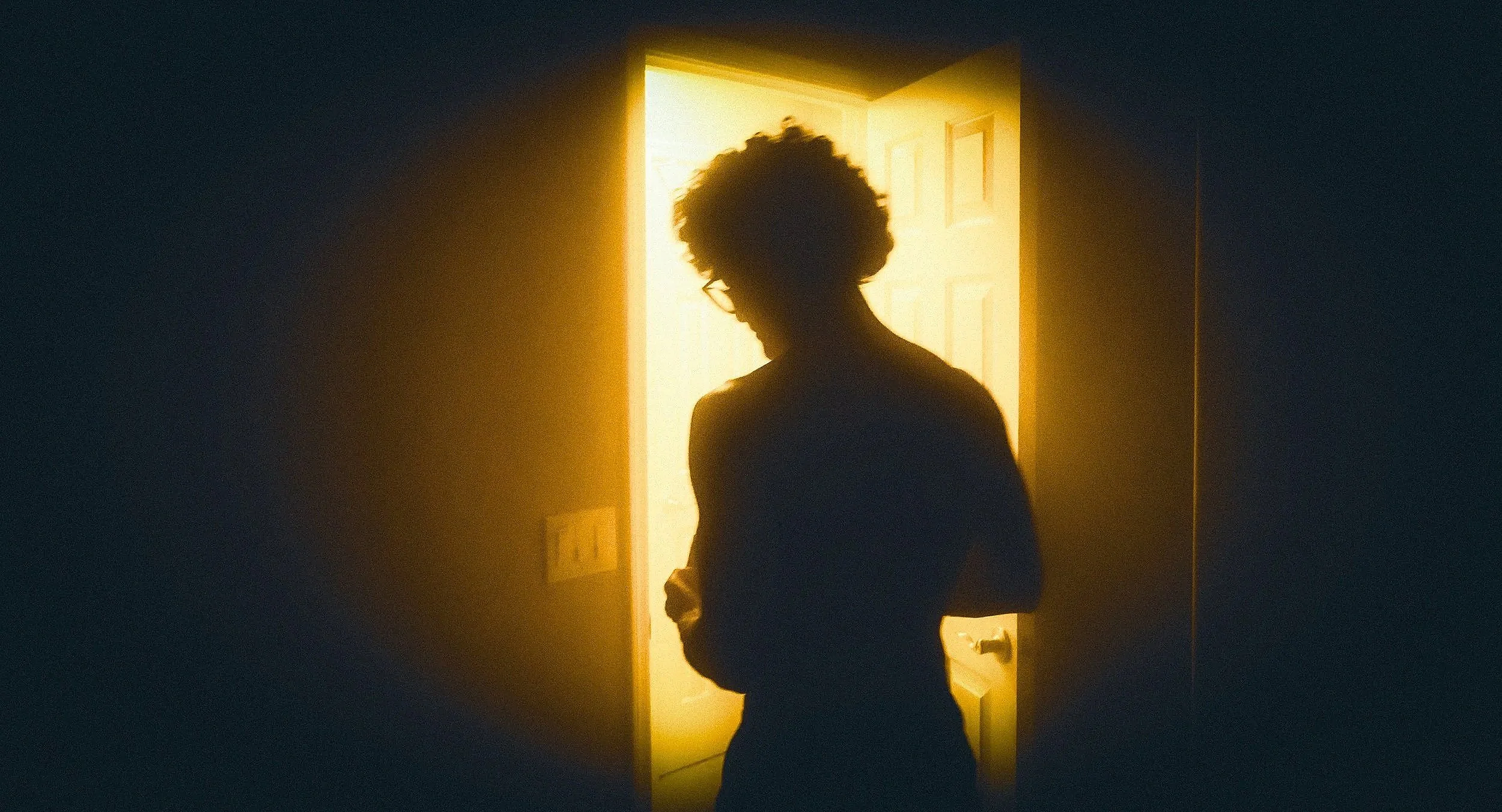 Silhouette of a person with curly hair and glasses standing in a doorway with bright sunlight coming through.