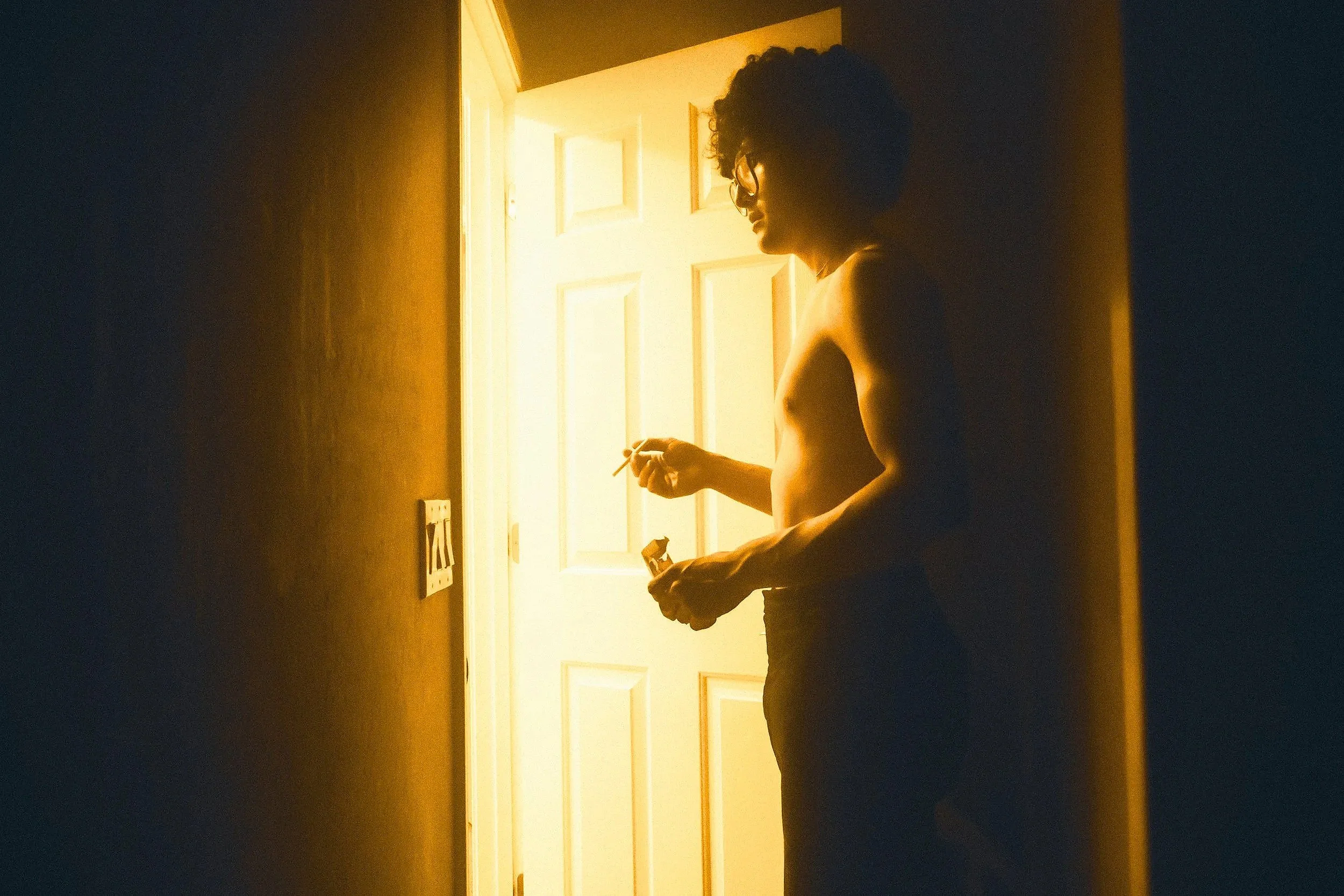 A shirtless man with curly hair and glasses standing inside a dim room near a door with light shining from behind, holding a cigarette and a small object in his hands.